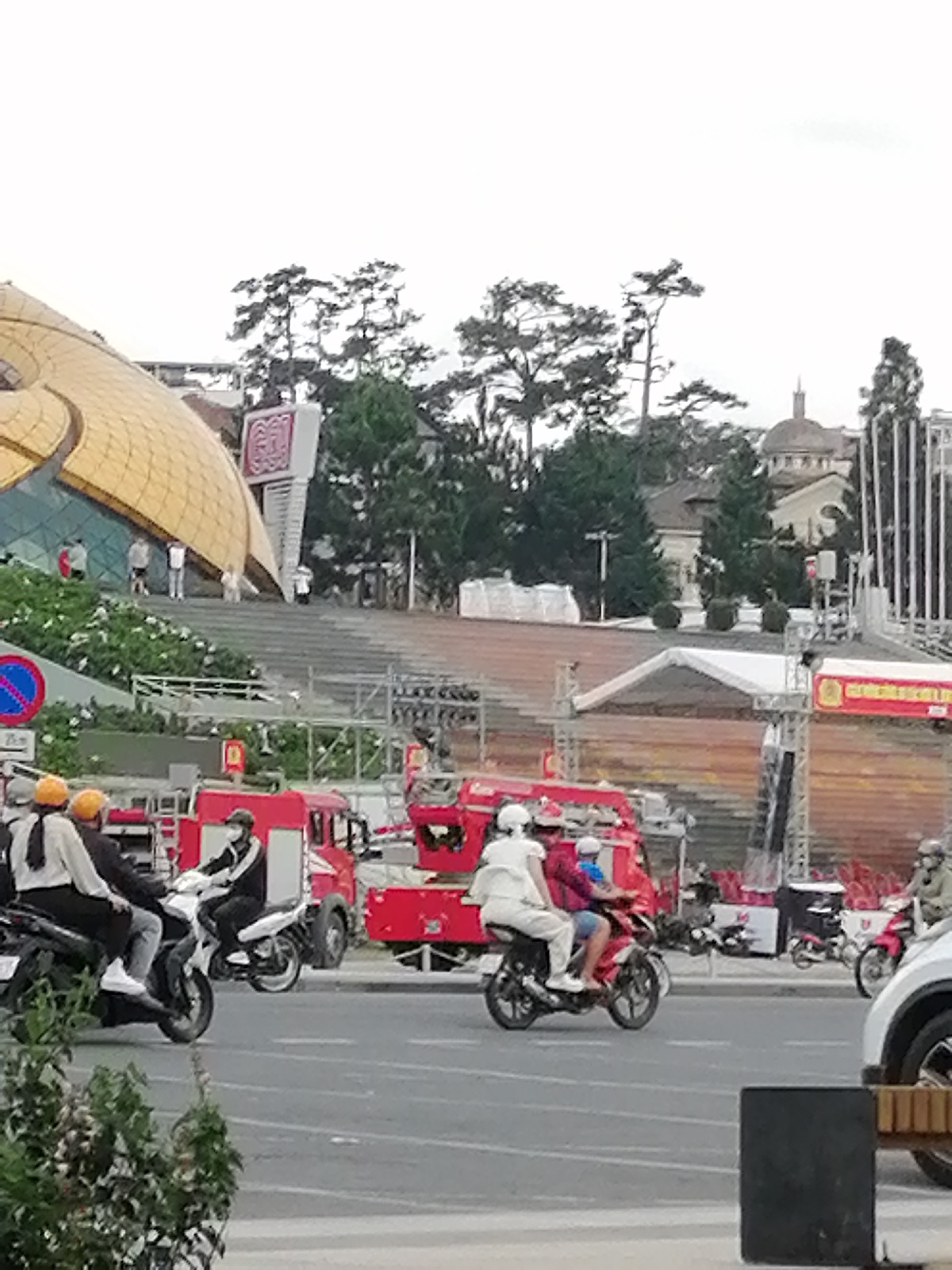 In Da Lat, Vietnam, on a street in front of a modern building with a distinctive golden dome, a busy scene unfolds. Several motorbikes are in motion, with riders and passengers navigating the road. In the foreground, a group of individuals on motorbikes are visible. One motorbike carries two people; the rider wears a white shirt and helmet, while the passenger is in a white outfit. Another motorbike nearby has a rider in a white shirt and black pants, wearing a helmet. Further back, a large red fire truck is parked, alongside other vehicles. The background features lush green trees and a building with a domed roof, characteristic of the city's architecture. The overall atmosphere suggests a vibrant urban environment with ongoing activity.