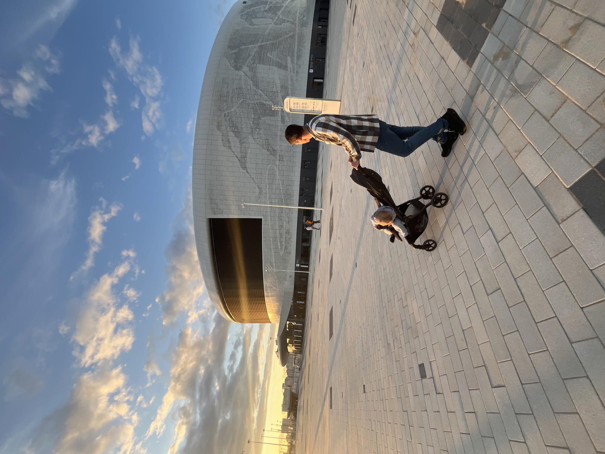 In the city of Nizhny Novgorod, Russia, an individual pushes a stroller across a large, paved plaza in front of a modern, curved stadium with a white, perforated facade. The sun is low in the sky, casting long shadows and illuminating the clouds with warm, golden light. The stadium features a dark, reflective strip near its top, and a large screen is visible within. The paving stones are light gray and laid in a rectangular pattern. Several other individuals are visible in the distance, near the entrance of the stadium. The scene captures a peaceful moment outdoors, likely in the late afternoon or early evening.