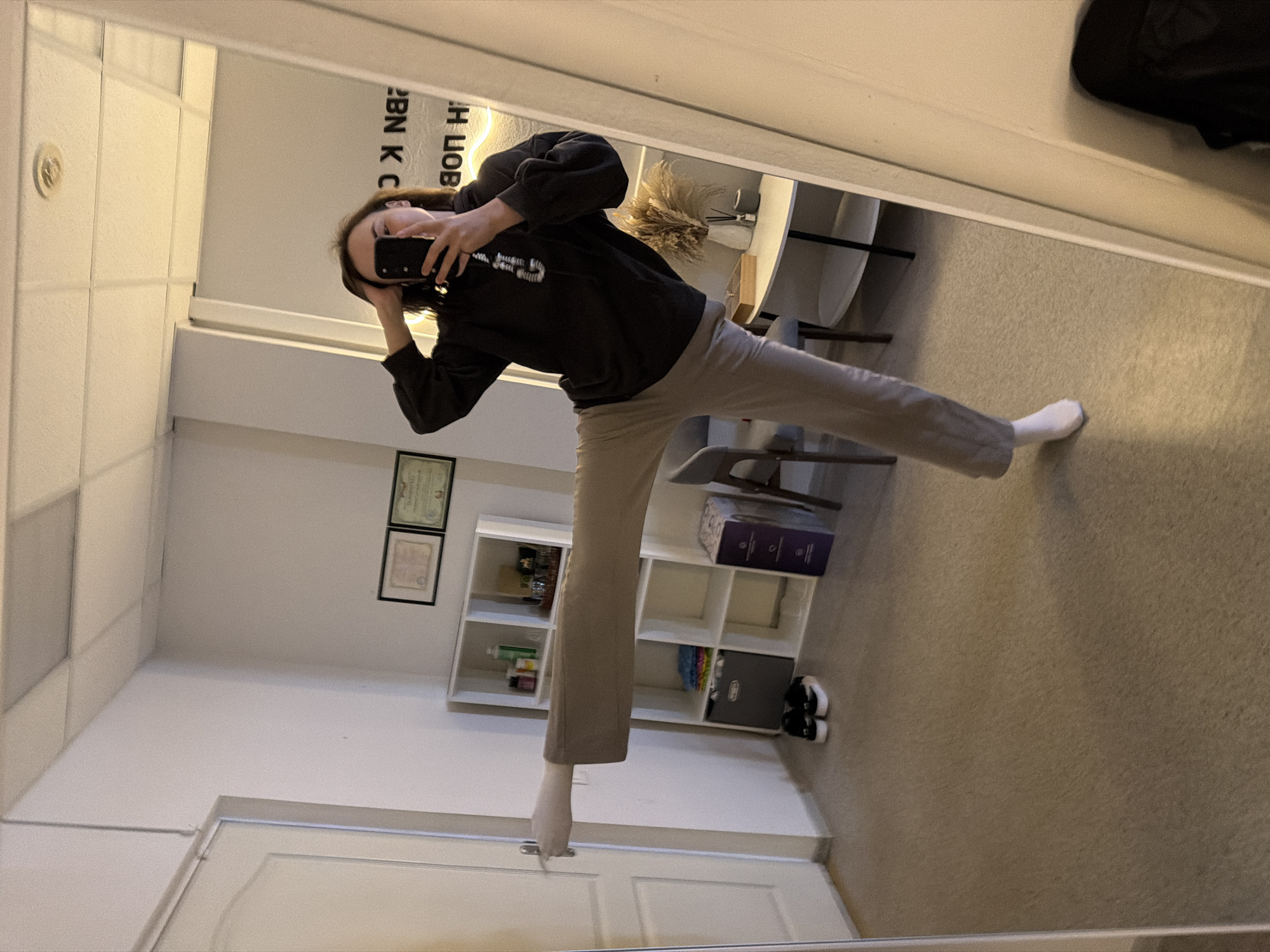 A woman is taking a selfie in a mirror. She is wearing a dark hoodie and light-colored pants. One foot is on the carpet, and the other is near a white door. The background includes a bookshelf with various items, two framed certificates on the wall, and a small table with decorative plants. The room appears to be in a residential setting. It is not possible to confirm the location in the city of Chaikovsky, Russia from the provided image.