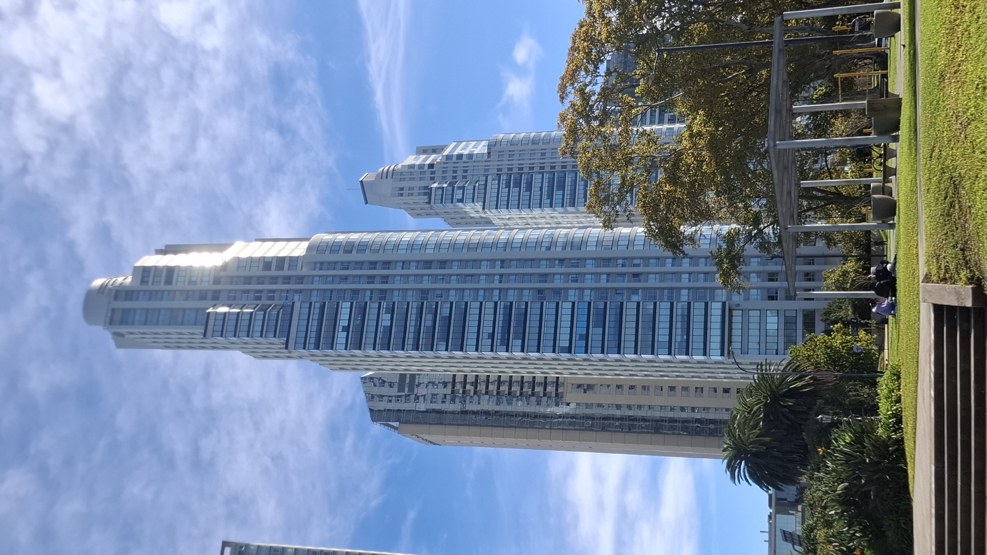 The image shows three residential towers in Buenos Aires, Argentina, near the river. They are partially obstructed by greenery. The weather is sunny with a blue sky and scattered clouds. A park area with trees, benches, and some people sitting is visible in the foreground. There is also a stone structure.