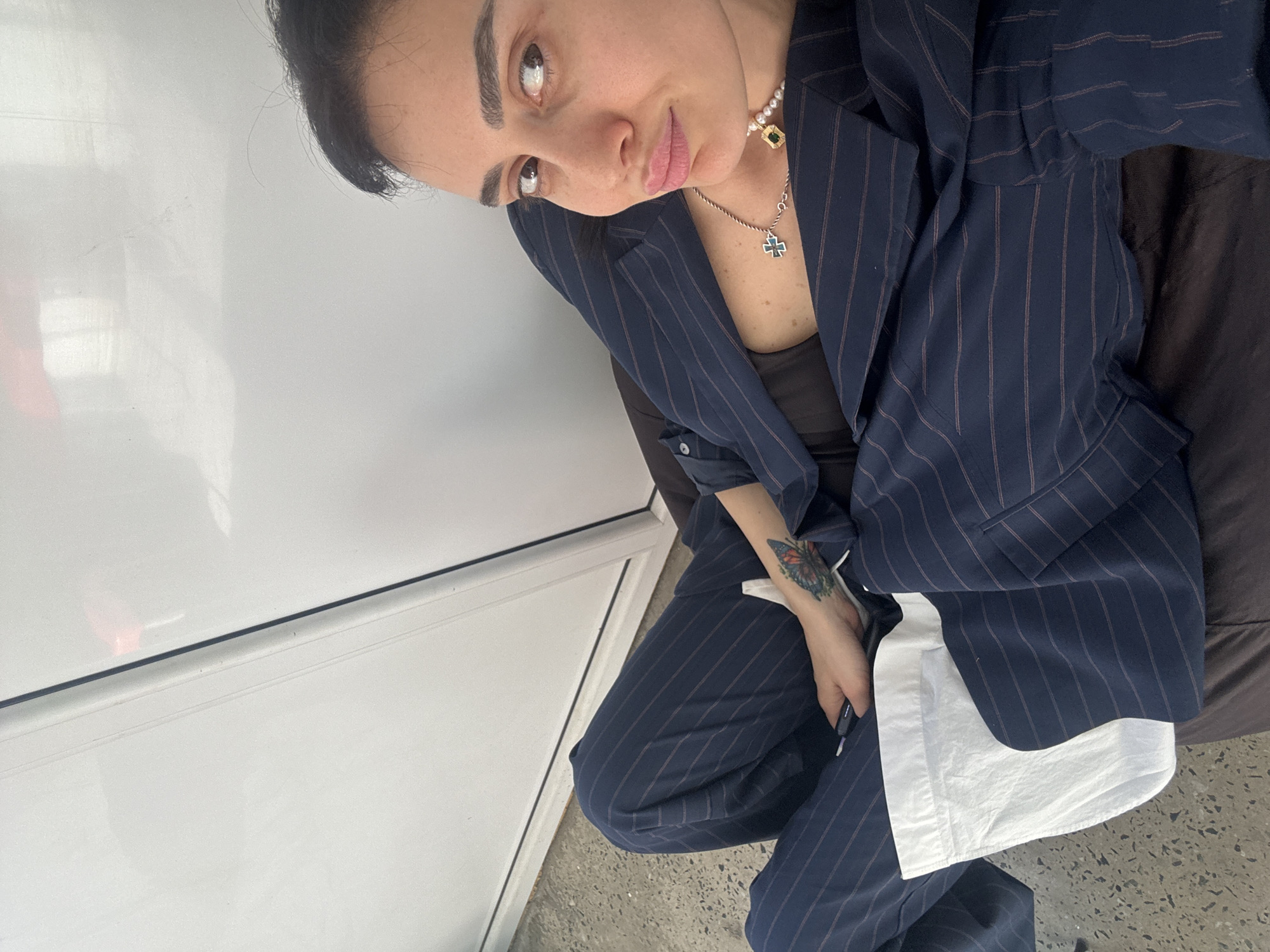 A young woman with dark hair pulled back poses for a photo, looking upwards and to the camera. She has full eyebrows and her lips are slightly parted in a natural pout. She is wearing a navy blue pinstripe suit jacket, which is open, over a dark, possibly black, top. Matching pinstripe trousers are also visible.
Around her neck, she wears two necklaces: a pearl choker with a small gold-set, rectangular green gemstone pendant, and a longer silver chain with a small blue and white cross pendant. On her left forearm, a vibrant, colorful butterfly tattoo is visible. Her right hand is resting on her leg, holding a slim, dark object, possibly a phone or pen, and some white fabric, resembling a shirt cuff, is visible around her wrist.
She appears to be sitting in an indoor setting. To her left is a plain white wall or panel with a subtle horizontal seam. To her right and behind her is a dark brown, possibly upholstered, surface. The floor beneath her is light-colored and speckled, suggestive of concrete or terrazzo. The lighting is bright and even, indicating an indoor environment. There is no visible text in the image.