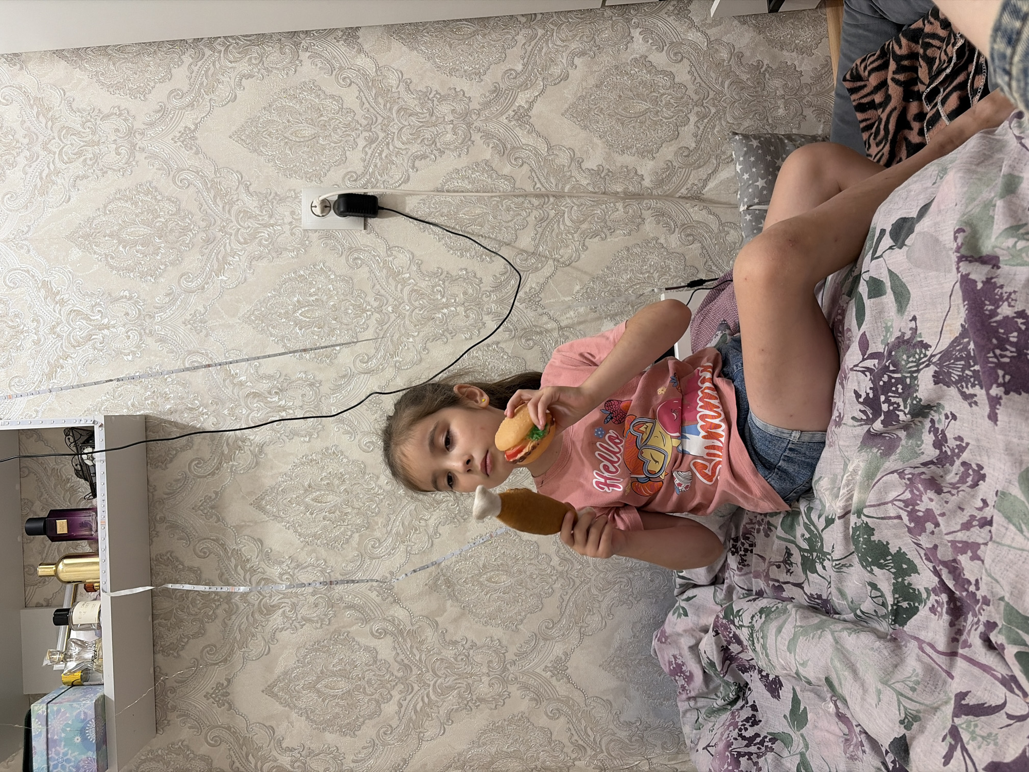 A young girl sits on a bed covered with a floral patterned comforter, holding a toy chicken leg in her left hand and a toy hamburger in her right hand. She wears a pink t-shirt with cartoon characters and denim shorts. Behind her, a wall is covered with ornate, light-colored wallpaper. A white shelf is mounted on the wall above the bed, displaying several perfume bottles and a small blue box. An electrical outlet with a black adapter is visible on the wall. The scene is indoors, likely a bedroom, and the lighting suggests it is daytime. There is no indication of the location being Tambov, Russia.