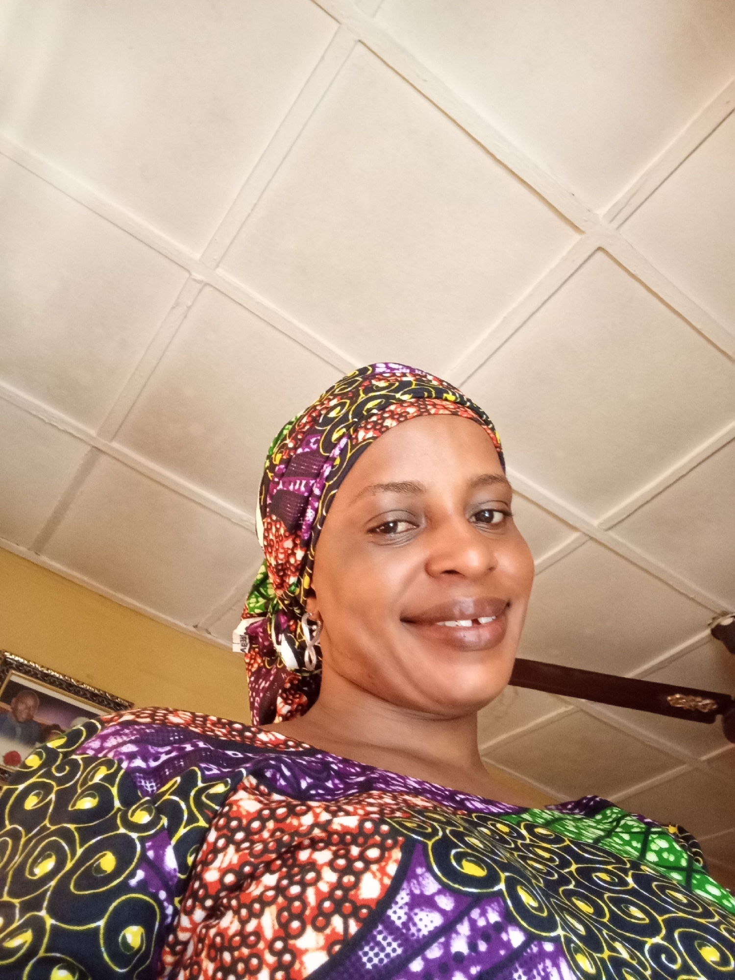 A woman wearing a colorful headscarf and a patterned top is looking directly at the camera, smiling. The background features a white, tiled ceiling and a hint of a yellow wall. A ceiling fan is partially visible in the upper right corner. The overall scene suggests an indoor setting, likely a home in Narayi, Nigeria, as specified. A framed photograph can be seen on the wall in the bottom left corner.