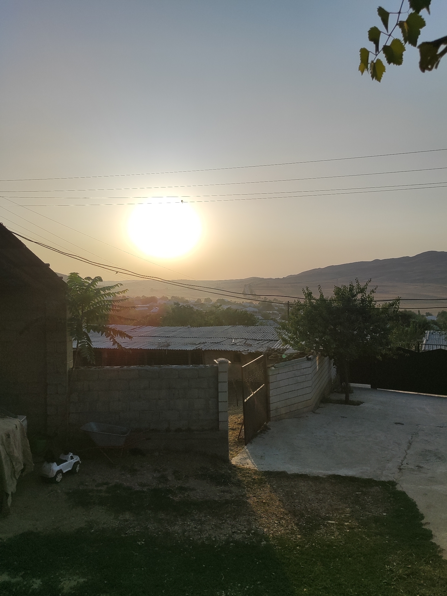 The image depicts a rural scene at dusk in Sultonobod, Tajikistan. The setting sun casts a warm glow across a landscape of low-lying buildings with corrugated metal roofs, trees, and distant hills. In the foreground, a partially constructed brick wall divides a yard. To the left, a dark-colored house with a sloped roof is visible, along with a tarp-covered object. A child's white toy truck and a wheelbarrow are present on the ground. Several power lines crisscross the sky above the scene. A few leaves from a tree are in the upper right corner of the frame.