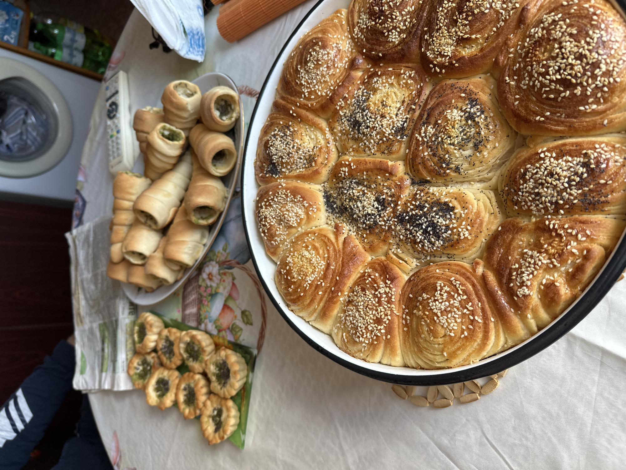 The image depicts a table laden with baked goods. A large, round baking dish filled with golden-brown, spiraled rolls sprinkled with sesame seeds and poppy seeds dominates the right side of the frame. To the left, a plate piled high with pastry cones, filled with a greenish filling, and a small rectangular dish holding round pastries with similar filling are visible. A washing machine and assorted items are visible in the background, suggesting a domestic kitchen setting. The image was likely taken indoors, showing a home-baked spread. The city or country is not discernible from the image.
