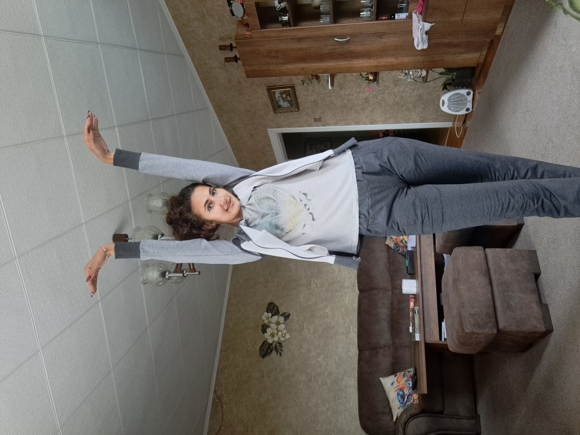 A woman with curly hair is lying on her side in what appears to be a living room. She is wearing a grey sweatshirt and pants. Her arms are extended straight out to either side. The room contains a brown couch, a wooden cabinet with glass doors, and a decorative flower arrangement on the wall. The ceiling has a textured, tiled pattern. The lighting is bright.