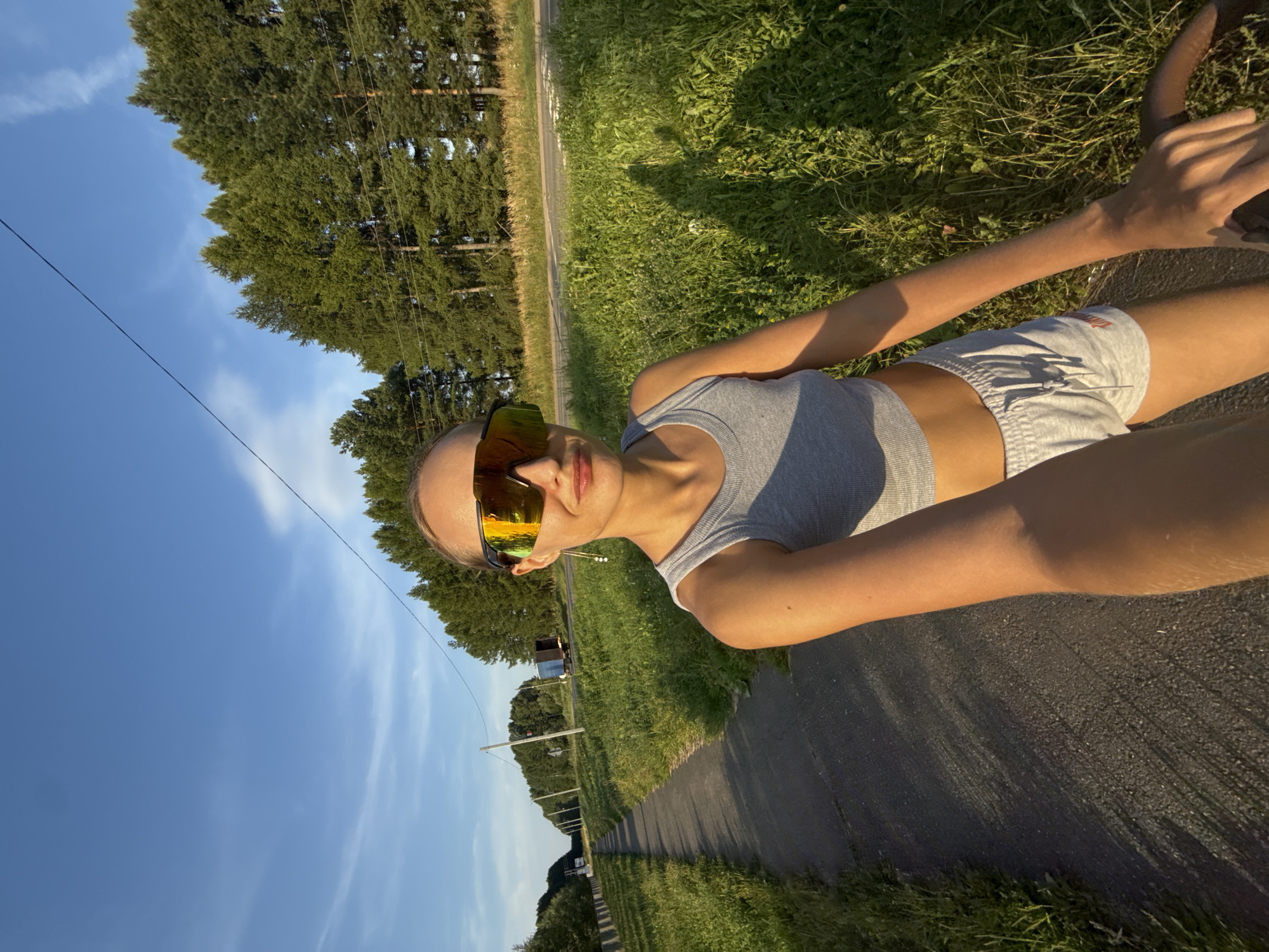 A selfie captures a young woman wearing sunglasses and a grey crop top and white shorts. She stands on a paved path next to a grassy area and a road. Tall pine trees line the right side of the road, and the sky above is blue with wispy clouds. The setting appears to be Podshivalovo, Russia.