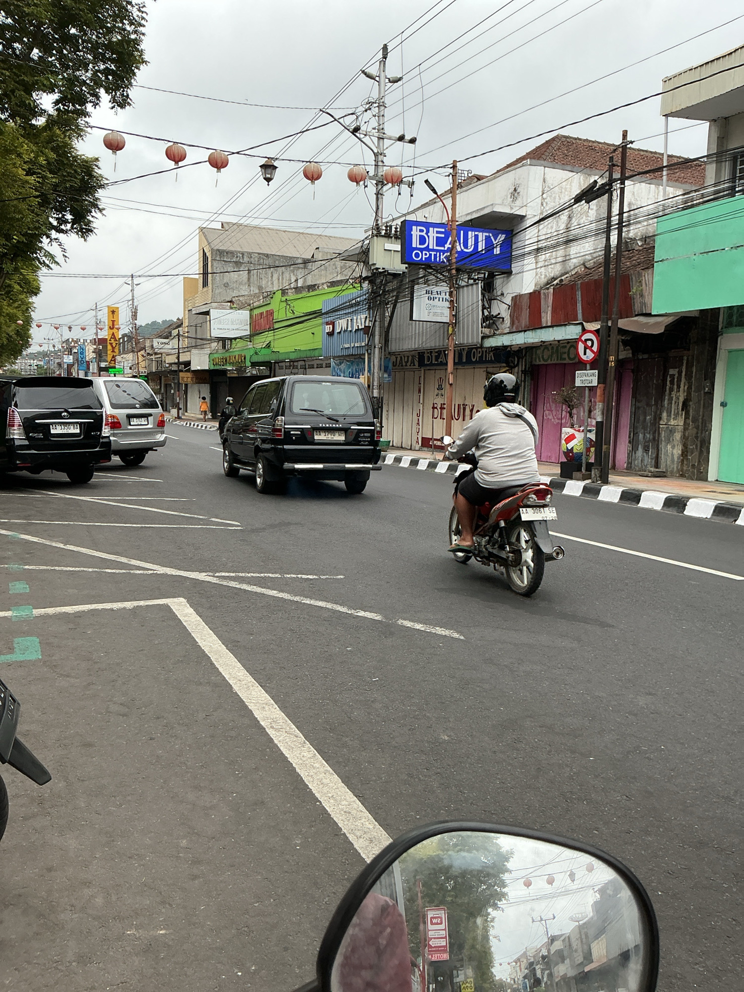 The image depicts a street scene in Magelang, Indonesia. The perspective is from a vehicle, possibly a motorcycle, looking forward and slightly to the right. In the foreground, a motorcycle is being ridden by a person wearing a gray t-shirt, black shorts, a black helmet, and flip-flops. The motorcycle is red and silver, with the license plate "AA 3061 SE".
The street is paved and marked with white lines. Several cars are also present, including two black SUVs and a silver SUV. The buildings lining the street are a mix of styles, with some appearing older and others more modern. Signs are visible on the buildings, including one that reads "BEAUTY OPTIK" in blue and white lettering. Red lanterns are strung across the street, suggesting a festive or cultural event. The sky is overcast.
In the background, a street sign with a red circle and a diagonal line indicates a prohibition, likely a "No U-turn" or similar traffic regulation. A small white sign below it reads "DISEPEMIANNG TROTOAR," which translates to "Sidewalk reservation." The overall atmosphere is that of a typical urban street in Indonesia, with a blend of traffic, commerce, and pedestrian activity.