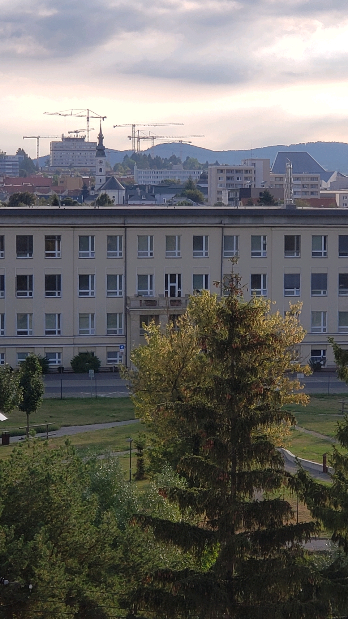 The image captures a view of a large, cream-colored building with numerous windows, likely a hospital or institutional building, in Prešov, Slovakia. Several trees are in the foreground, with dense evergreen foliage in the lower right and a sparser, sunlit deciduous tree to the right of the center. Beyond the cream building, the cityscape of Prešov is visible, featuring other buildings, a church steeple, and multiple construction cranes dominating the skyline in the distance. The sky is overcast with some lighter patches. The scene is a wide shot, focusing on the architecture and the surrounding urban and natural elements under a cloudy sky.