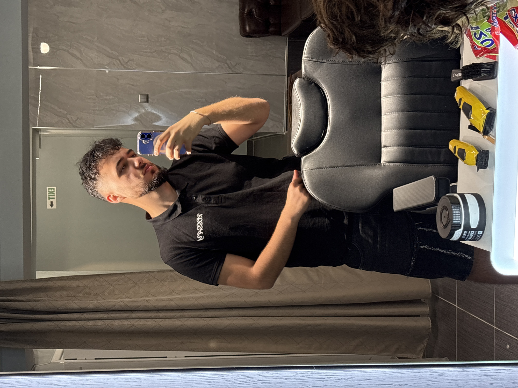 A man is captured in a mirror selfie within an indoor setting, likely a barbershop or salon in Varna, Bulgaria. He is visible from the chest up, with his head tilted slightly, focused on the blue-cased smartphone held in his right hand. He has short, dark curly hair and a well-trimmed beard. He is wearing a black short-sleeved polo shirt with a white logo, possibly "UNXOR," and black shorts.
To his right, a black leather salon or barber chair with padded sections is prominently featured. On a white counter or shelf adjacent to the chair, various grooming tools and items are arranged: two distinct yellow hair clippers/trimmers, a black brush, and a red and white bag of snacks with the number "50" and partial text, possibly "JAGUAR." A white cylindrical bottle with a black cap, bearing labels like "CHEM" and "WWW.OPI," is also present.
The background reflected in the mirror includes a grey tiled wall above the man's head, and below him, a patterned brown curtain and a white air conditioning unit. A reversed "EXIT" sign is visible on a grey wall section to his left. The mirror's bottom edge is framed by a wooden-textured surface. The lighting is internal, offering no clues about the time of day or outdoor weather. The man's expression is neutral and concentrated on his phone.