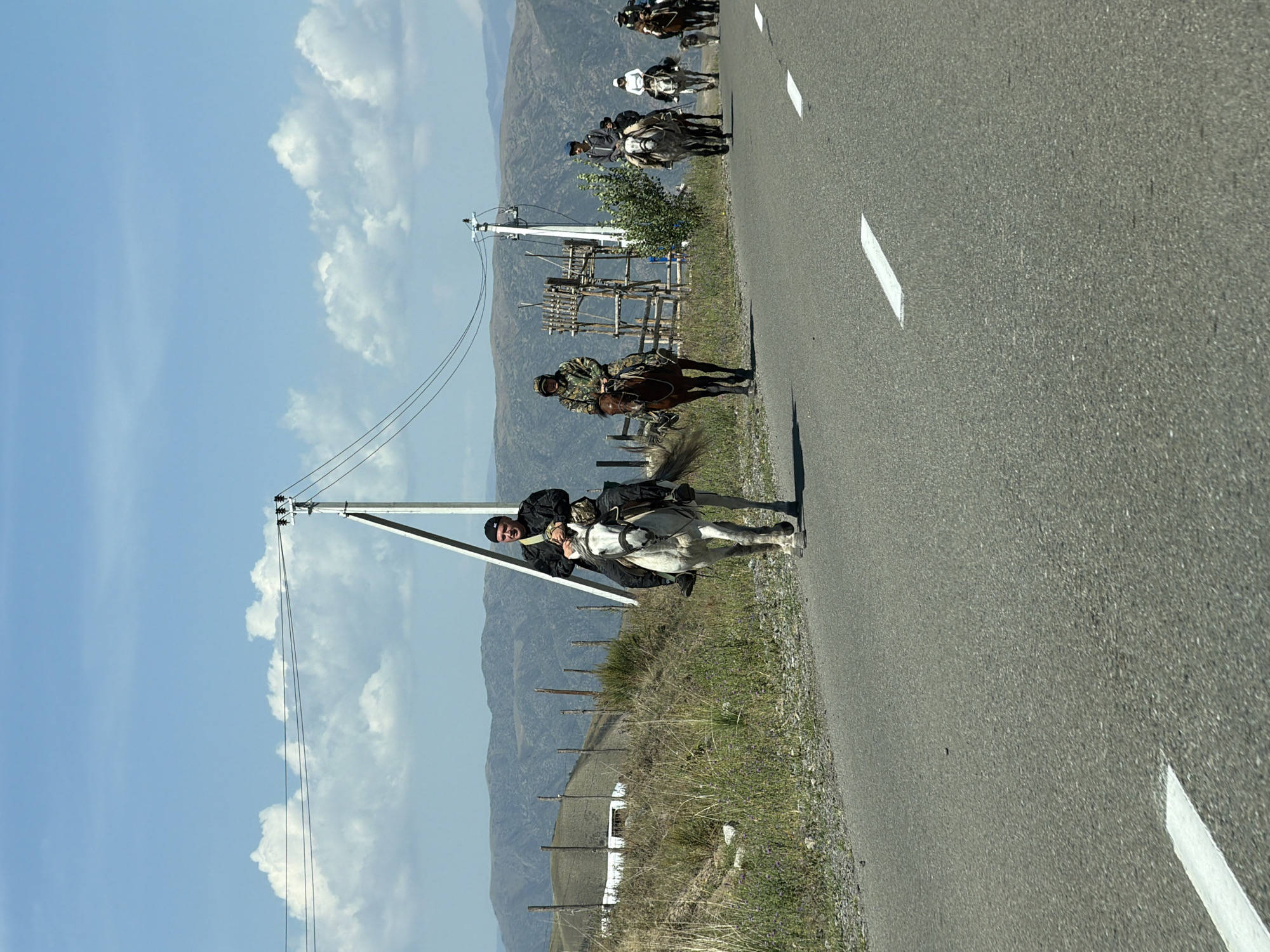 In the city of Kontora Kazlesa, Kazakhstan, a group of individuals are riding horses on an asphalt road. The sky is blue with scattered clouds, and rolling hills form the background. The riders are dressed in various attire, some in traditional clothing and others in more modern outfits. The horses are of different colors and breeds, and they appear to be walking or trotting along the road. A utility pole with power lines stands on the side of the road, and there are some buildings and fences visible in the distance. The overall scene suggests a rural or semi-rural setting, possibly during a parade or a cultural event.