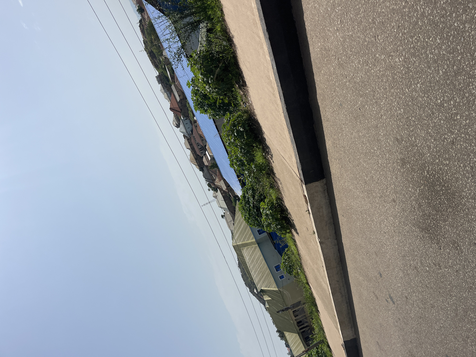 The image shows a street scene with several houses and vegetation in the city of Gura Top, Nigeria. In the foreground, a paved road runs diagonally across the bottom of the frame, bordered by a concrete curb with black and white markings. Beyond the curb, a sidewalk area with some patchy grass and dirt is visible.
Several buildings are present in the background, situated on a gentle incline. One prominent house has a light yellow, corrugated metal roof. To the right of this, another building features a bright blue corrugated metal roof. Further up the slope, a cluster of houses with various colored roofs, including red and brown, are visible. The houses appear to be residential structures.
The environment is sunny with a clear sky, suggesting a daytime setting. Some power lines are strung across the upper portion of the image, running diagonally. Dense green vegetation, consisting of shrubs and bushes, is interspersed among the buildings. A large green plant is prominently featured in the mid-ground, obscuring some of the structures. The overall impression is a developing urban or suburban area with a mix of housing and natural elements. There are no people visible in the image.