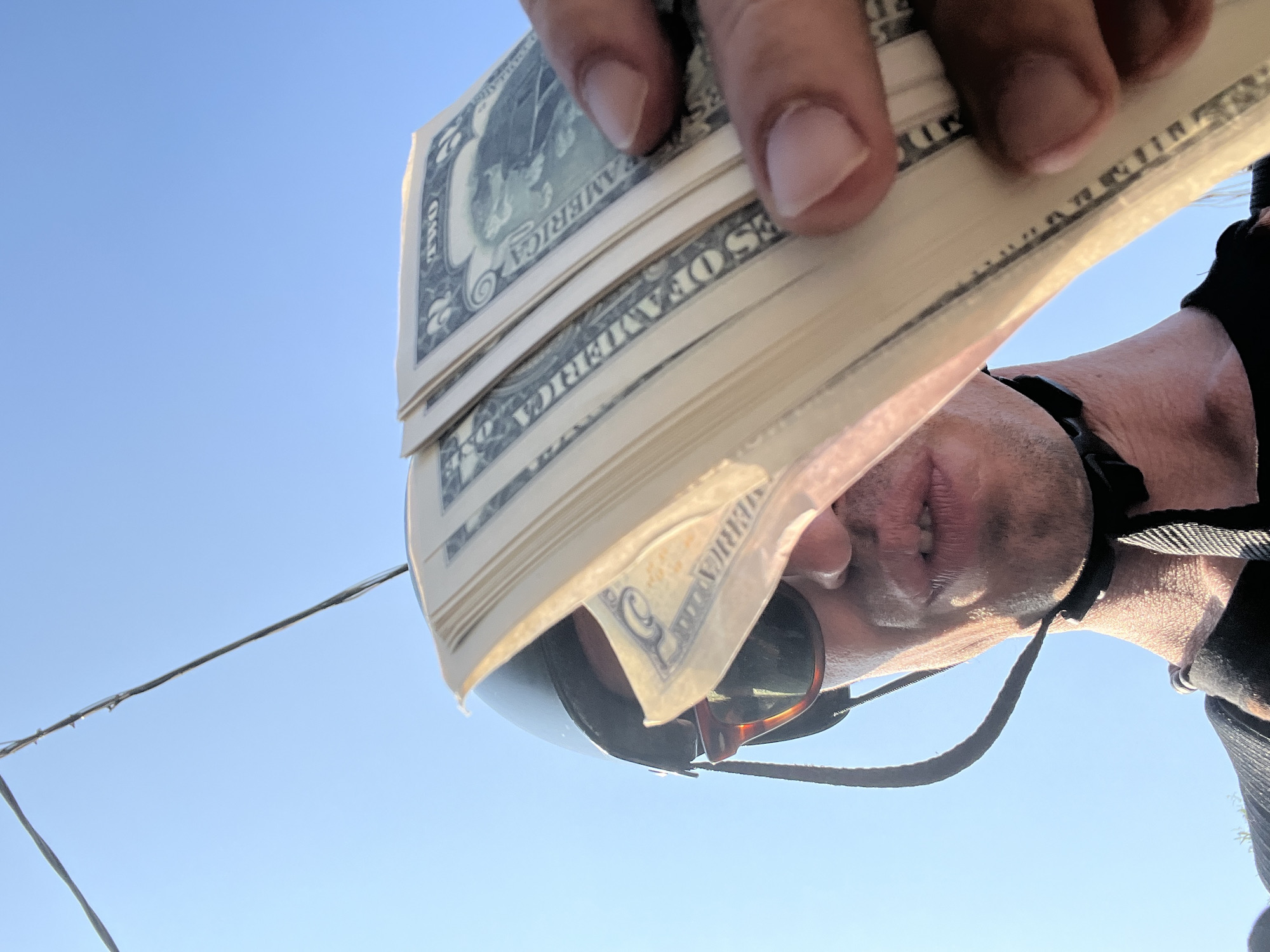 The image is a close-up, low-angle shot of a person holding a fan of US dollar bills in front of their face. The person is wearing sunglasses and a helmet with a chin strap. The background is a clear blue sky. The dollar bills appear to be two-dollar bills, with "TWO" visible on some of them. The person's lips are parted, and they are looking towards the camera. The lighting suggests it is daytime, possibly outdoors. There is no indication of the location being in the city of Mussolini, Italy, or any other specific city.