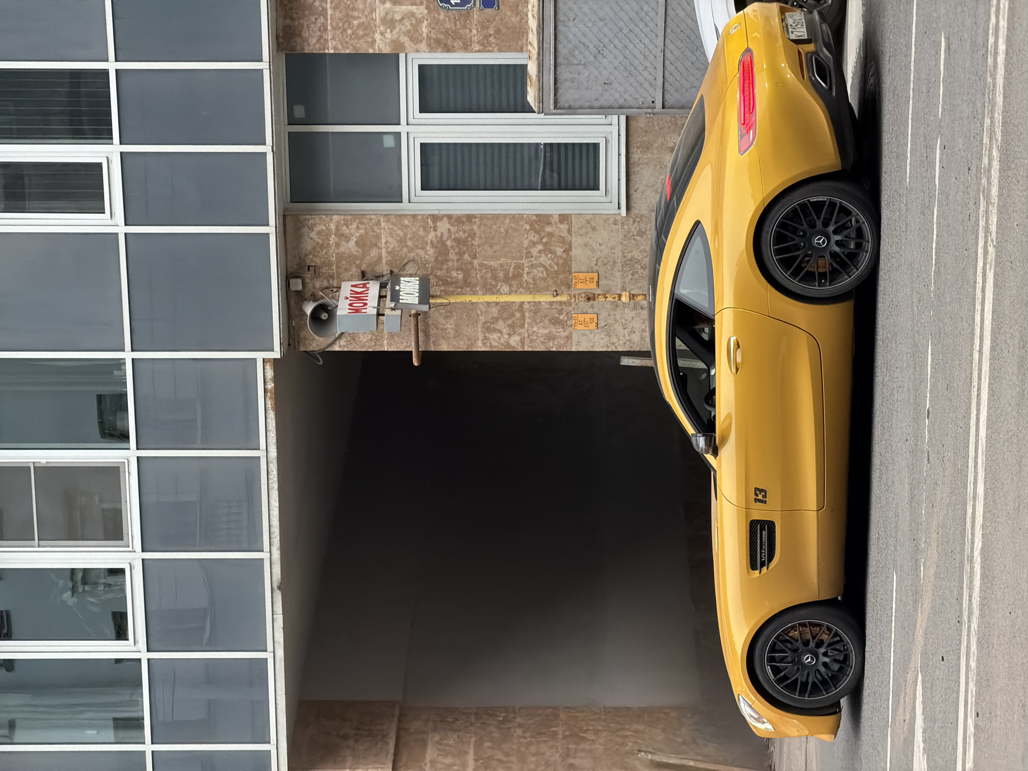 The image shows a bright yellow Mercedes-AMG GT parked on the side of a road. The car features a black "13" decal on the side and black wheels with Mercedes-Benz center caps. The rear license plate is partially visible. The location is indicated as Saint Petersburg, Russia.
The car is parked next to a building with a facade that includes a section with a grid of square windows and a section with a smaller, double window. A dark tunnel-like opening is also present. A sign in Russian is attached to the building, with the word "мойка" (moyka) visible, which translates to "car wash." A small speaker is mounted next to the sign. Gas pipes and utility boxes are also visible on the building's exterior. The street surface is asphalt with white lane markings.