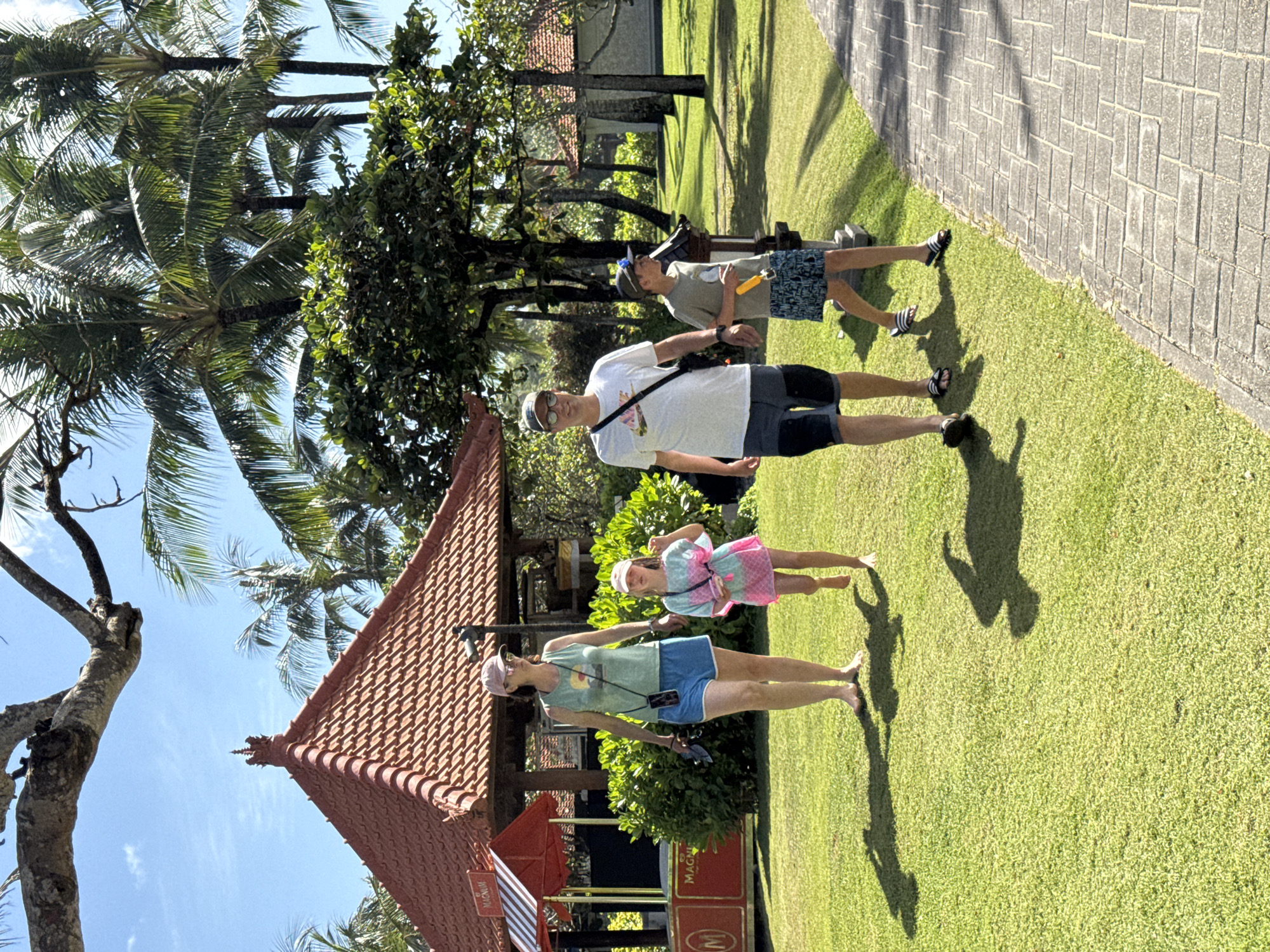 The image depicts a family of four standing on a grassy lawn next to a paved walkway. They are in the city of Kuta Selatan, Indonesia.
A woman in a t-shirt and shorts stands closest to a small building with a red tile roof. The building appears to be an ice cream stand with "Magnum" branding. A young girl in a dress stands next to her. A man in a white shirt and shorts stands behind them, and a teenage boy in a t-shirt and shorts stands further back. The background includes palm trees and other greenery.