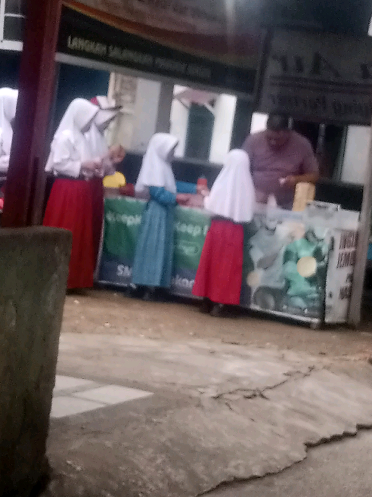The image depicts a street scene in Pekanbaru, Indonesia. Three young individuals, appearing to be female and dressed in white headscarves and long skirts of red and blue, are standing in front of a vendor stall. One is wearing a red skirt, another a blue skirt, and a third, only partially visible, also appears to be wearing a red skirt. A man, presumably the vendor, is visible in the background behind the stall, wearing a purple shirt. The stall itself is adorned with various advertisements and logos, including one with green lettering. The ground surface appears to be a mix of dirt and paved areas, with some cracked concrete visible in the foreground. The overall lighting suggests it is daytime, but the image quality is blurry, making fine details difficult to discern.