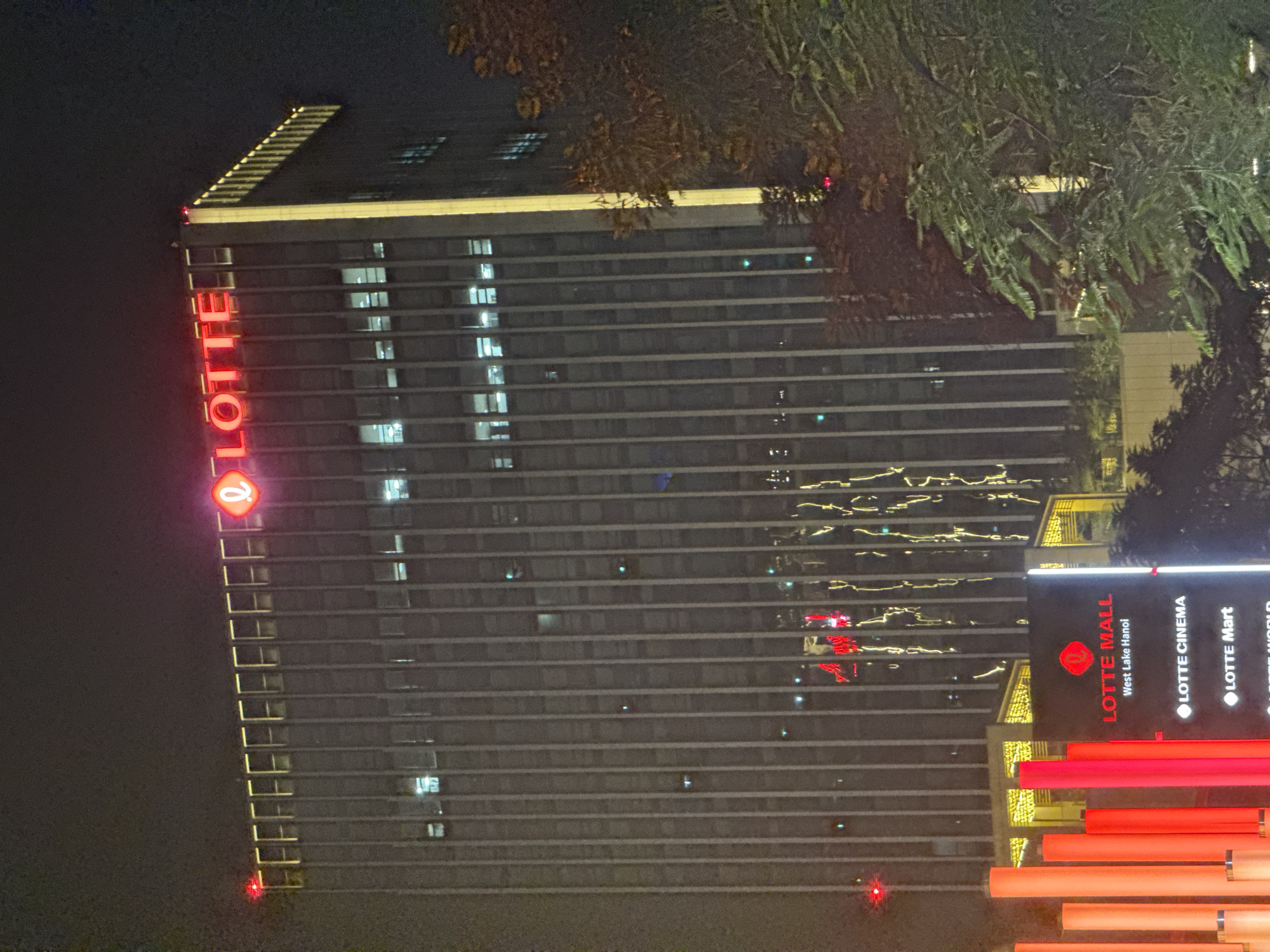 The image captures a nighttime view of the Lotte Mall and Lotte Center in Hà Nội, Vietnam. The Lotte Center, a towering skyscraper, is illuminated with the Lotte logo prominently displayed in red neon letters at its apex. The building's facade is lined with vertical elements and windows, some of which show interior lighting. Below the skyscraper, Lotte Mall is visible, with illuminated signage indicating "LOTTE MALL West Lake Hanoi," "LOTTE CINEMA," and "LOTTE Mart." The foreground features a cluster of tall, red, cylindrical structures, possibly part of an artistic installation or architectural feature. Trees with dense foliage are present on the right side of the frame, obscuring parts of the building and adding to the nighttime atmosphere. The scene suggests a bustling urban environment illuminated by artificial lights.