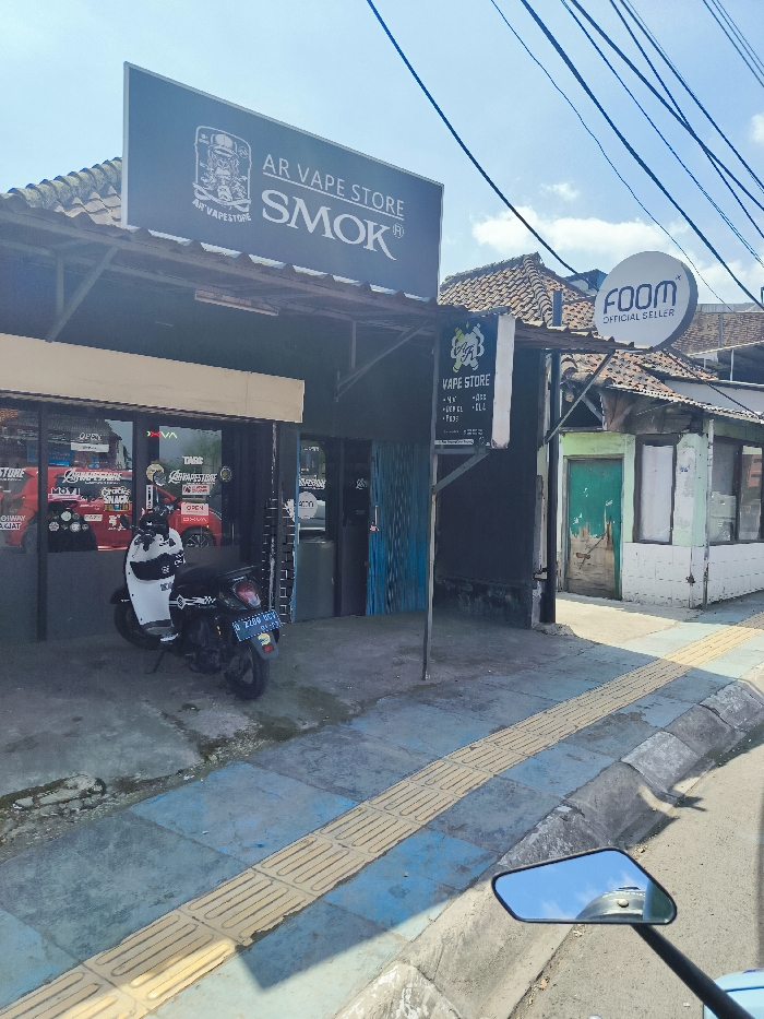 The image shows a street-level view of a storefront in Padalarang, Indonesia, located at latitude: -6.851, longitude: 107.497. The storefront has a black awning with a sign that reads "AR Vape Store Smok." The store appears to be a vape shop, with a sign advertising "Vape Store" and a sign with "Foom Official Seller." A motorcycle is parked in front of the store. The sidewalk is paved with concrete and yellow tiles. There is a curb separating the sidewalk from the street. The street is paved with asphalt. The building next door to the store is a simple, one-story structure with a green door and white walls. The sky is clear and blue. The sun is shining. A small motorcycle mirror reflects the street in the lower right corner of the image.