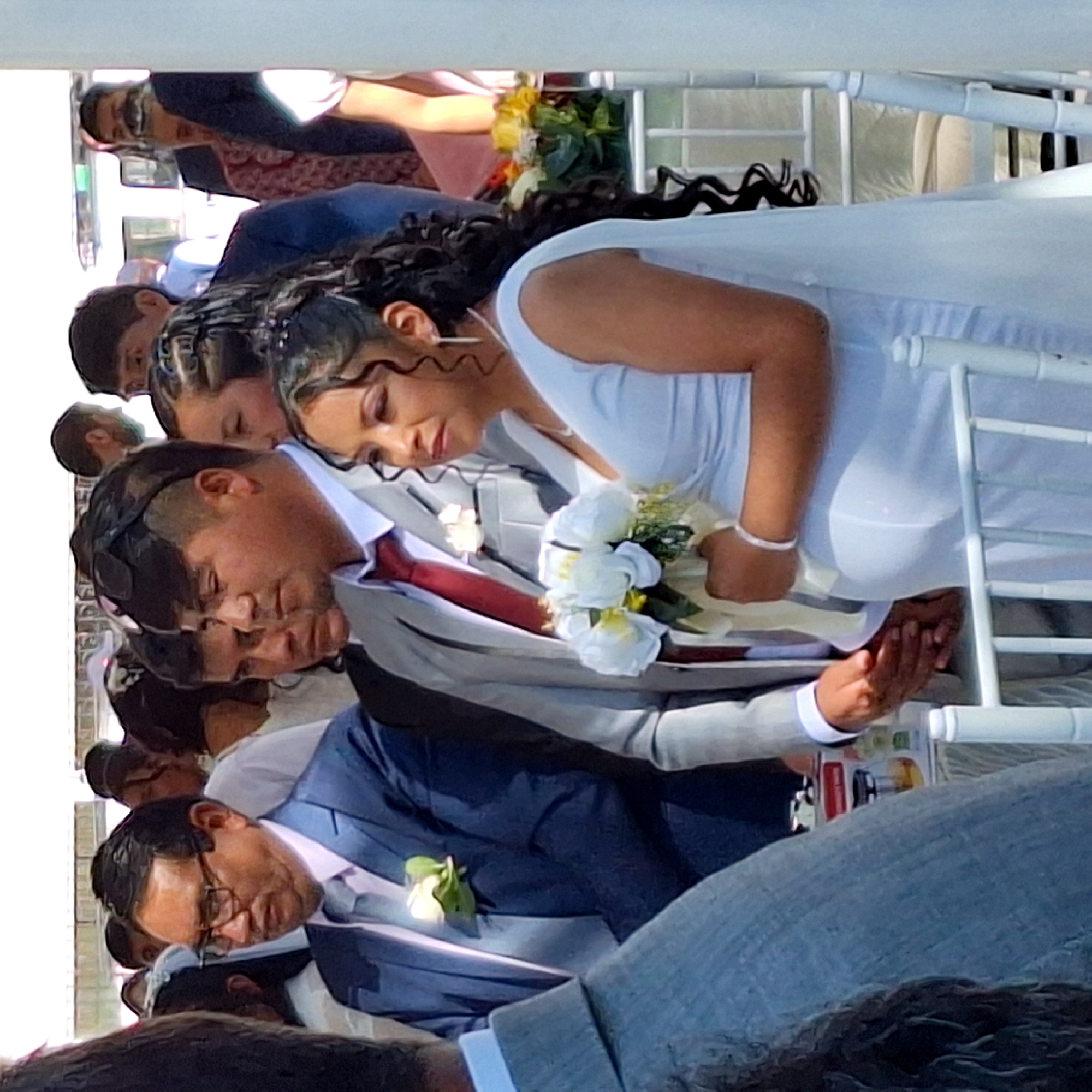 The image depicts a wedding ceremony, likely taking place in Cerro Colorado, Peru. A bride in a white dress is looking at the groom who is wearing a gray suit with a red tie and holding her hand. She is holding a white bouquet of flowers. They appear to be seated, perhaps on a white bench. Other attendees are visible in the background, dressed in suits and dresses, seated in rows. Some attendees are wearing sunglasses. The general setting suggests an outdoor venue, possibly with chairs set up for the guests.