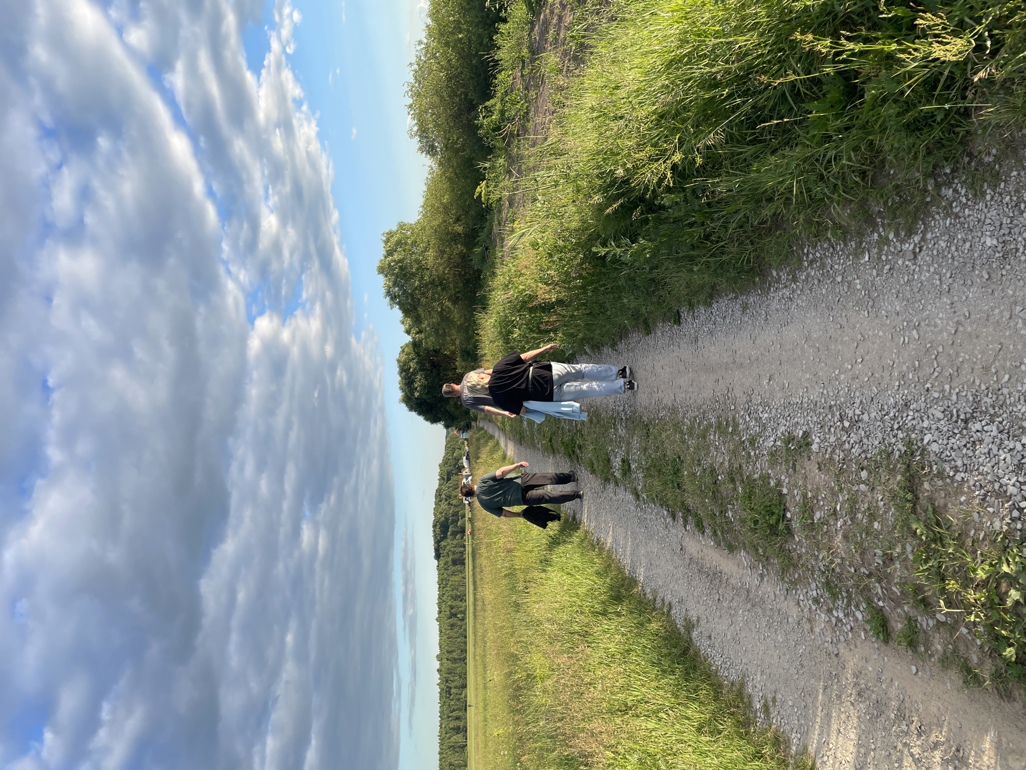Three individuals are walking away from the camera on a gravel path that runs through a grassy field. The person in the foreground, wearing a black shirt and light blue jeans, is a woman with blonde hair. To her left and slightly ahead is a person in a dark green t-shirt and dark pants, carrying a dark jacket. Further ahead, a person in a grey t-shirt is walking. The path is flanked by tall green grass. In the background, a dense line of trees forms a horizon, and beyond that, a village or settlement with buildings and parked vehicles is visible. The sky above is partly cloudy with patches of blue sky visible. The scene appears to be in a rural or semi-rural area, possibly on the outskirts of a settlement in the city of Турово, Russia, during daylight hours.