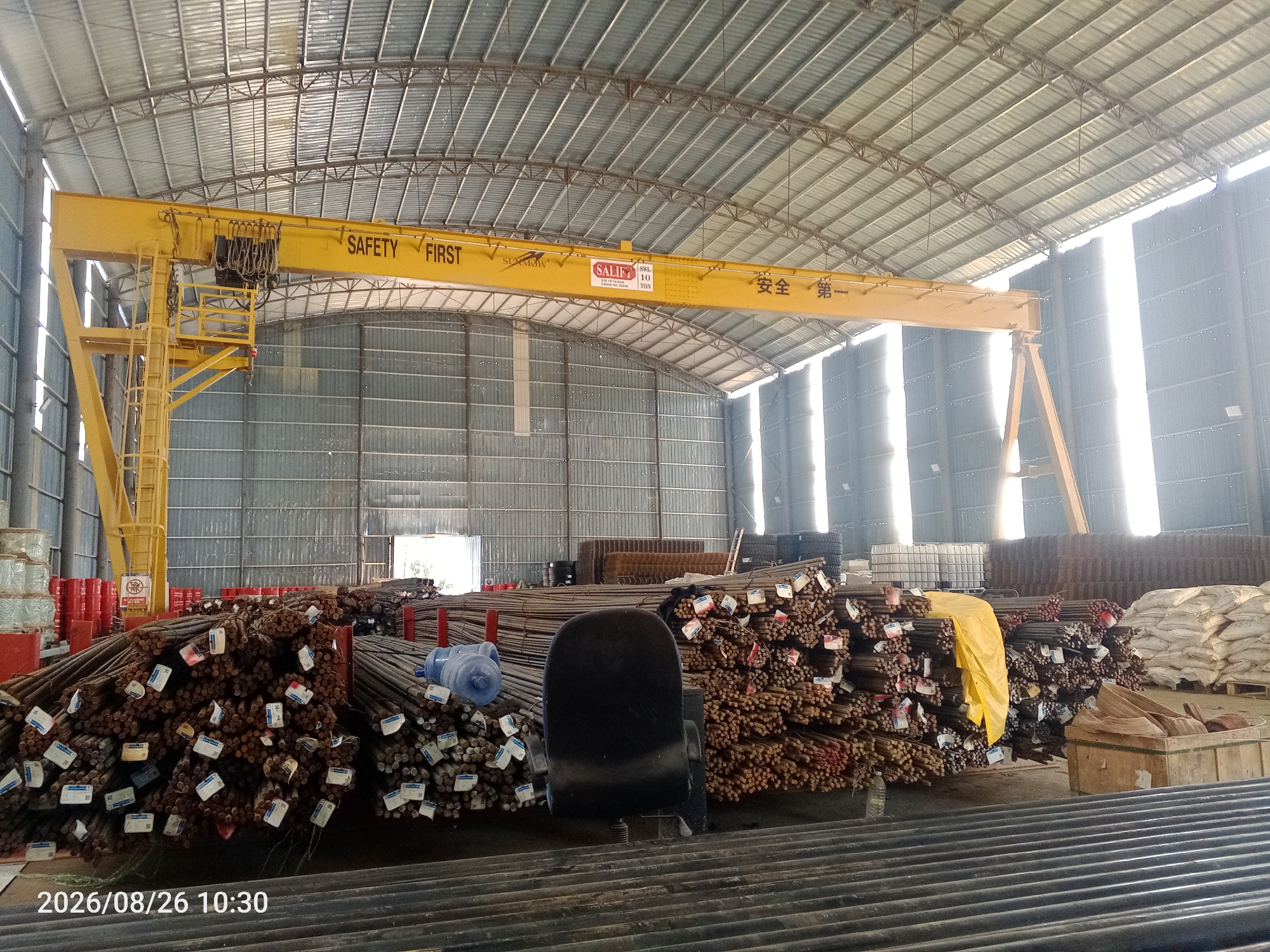 The image depicts the interior of a large warehouse in an industrial area of an unidentified city in Malaysia. The warehouse has a high, corrugated metal roof supported by a complex metal framework. Sunlight streams in from large openings on the right side of the building, illuminating the interior and creating bright patches on the walls and floor.
Dominating the scene is a yellow overhead gantry crane, labeled with "SAFETY FIRST" and some Chinese characters that translate to "Safety First". A manufacturer's plate on the crane indicates "SALIFE SWL 10 TON". The crane extends across the width of the warehouse.
The floor of the warehouse is piled high with bundles of steel rebar, stacked in various heights and arrangements. Many of the bundles have tags attached to them, likely indicating specifications or identification. In the foreground, more rebar is visible, some appearing to be larger diameter pipes or conduits.
To the left of the image, there are stacks of red cylindrical objects, possibly drums, and some spools of wire or cable. A dark office chair is positioned in the middle of the rebar piles. On the right side of the warehouse, there are large white bags, possibly containing cement or other construction materials, and wooden crates. The walls of the warehouse are constructed from corrugated metal siding. The overall impression is of a busy storage or construction materials facility. The date stamp "2026/08/26 10:30" is visible in the bottom left corner of the image.