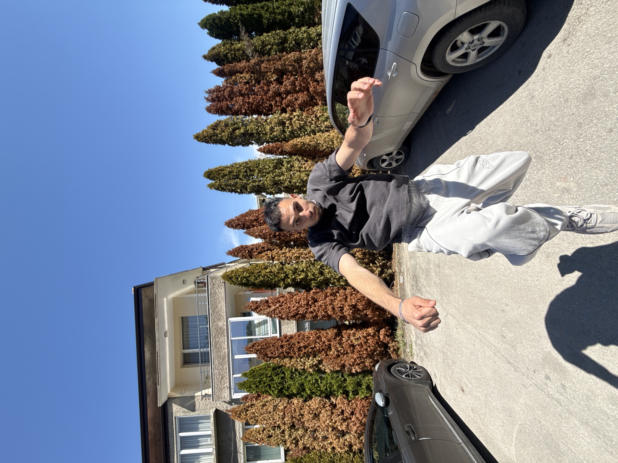 An outdoor, full-body portrait captures a young man posing on a sunny day in what appears to be a residential street in Shumen, Bulgaria. The image is oriented vertically with the man standing upright, looking directly at the camera with a neutral expression. He has short dark hair and is wearing a dark gray long-sleeved shirt, light gray sweatpants, and light gray sneakers. A silver chain bracelet is visible on his left wrist. His arms are slightly bent, with his hands held up near his waist.
In the background, a row of tall, conical evergreen trees, some appearing green and others a reddish-brown, line the street. Behind the trees on the left is a multi-story building with a light-colored facade, balconies, and multiple windows. Two cars are visible on the street: a light silver hatchback/SUV is parked on the right, partially visible behind the man, and a dark gray or black sedan is partially visible on the far left.
The time of day is bright, likely midday or early afternoon, as evidenced by the clear blue sky and the strong, distinct shadows cast on the asphalt or concrete street surface, including the man's shadow. The weather is clear and sunny. On the man's left pant leg, near the knee, light-colored, stylized letters, possibly "SOS," are visible. The overall scene suggests a casual, everyday moment captured in a European urban or suburban setting.