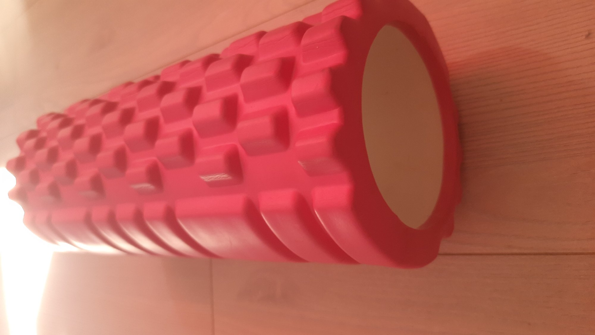 A bright pink foam roller with a textured surface rests on a wooden floor. The roller is positioned diagonally, with its wider end closer to the viewer and its textured surface facing upwards and away. The floorboards are visible beneath the roller and extend to the background, illuminated by a strong light source from the left, creating a bright, hazy area. There are no people or other objects visible in the image. The scene appears to be indoors, possibly in a home or gym setting. There is no indication of the city of Desnogorsk, Russia, in the provided image.