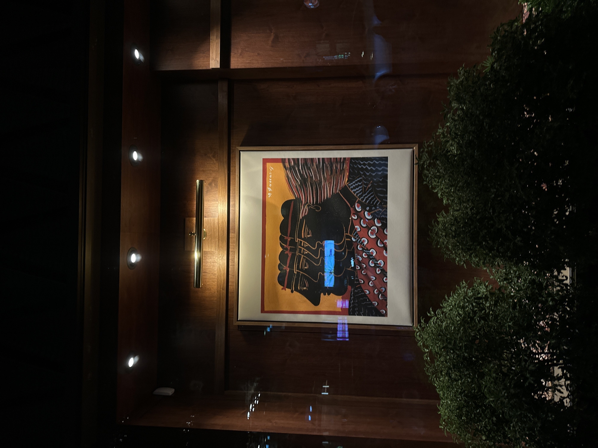 The image shows an interior scene with a focus on a framed artwork displayed on a dark wood paneled wall. The artwork depicts a profile of a person, rendered in a stylized and abstract manner with colors like yellow, black, and red. The wall is lit by small, recessed lights, and a brass-colored sconce is visible to the left of the artwork. Outside, seen through a likely glass wall or window, is foliage, possibly bushes or trees. Reflections are seen on the surface in front of the wall. Without more information, it is impossible to identify the exact location in London.