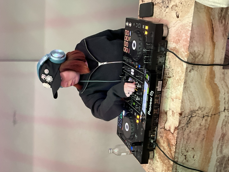 A young, fair-skinned woman with auburn hair is mixing music. She wears a black, zippered hoodie and a black cap adorned with jewel-like embellishments. Teal headphones rest atop her head. She manipulates controls on a Pioneer DJ system, positioned on a marble countertop. A black smartphone and a water bottle are nearby. The image is vertically oriented.