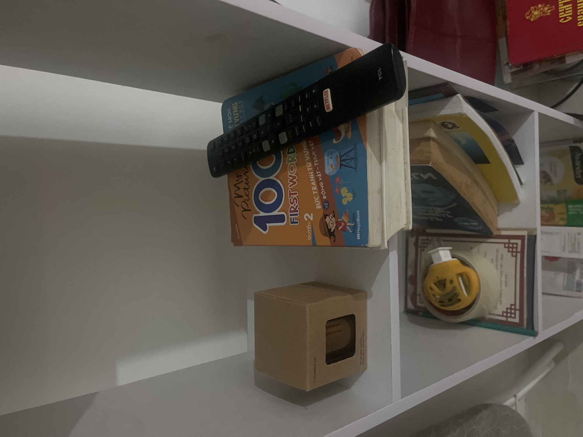 A black remote control with a Netflix button is lying on top of a stack of books. The top book is titled "My First 100 Words" and features illustrations of children and animals. Below it is another book with a blue cover. To the left of the books is a small, light brown cardboard box with a cutout window, revealing a yellow object inside. The scene is set on a white shelf, possibly in a residential setting in Nha Trang Ward, Vietnam, given the context of the question. The lighting is dim, creating shadows.