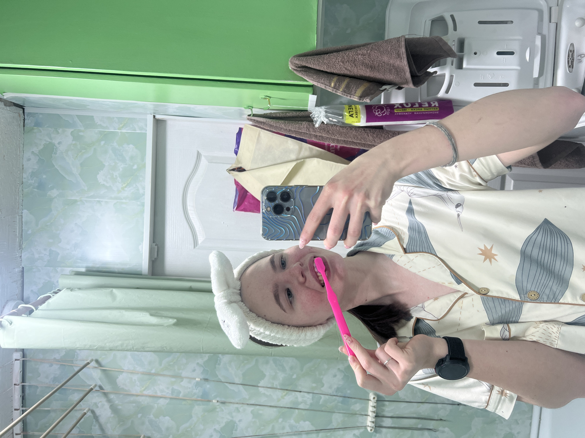 In a mirror selfie taken in a bathroom in Chaikovsky, Russia, a young woman is captured brushing her teeth. She appears to be in her late teens or early twenties, with dark hair pulled back by a white, fuzzy headband featuring bunny ears. She is wearing light-colored pajamas adorned with a pattern of grey-blue leaves and golden starbursts.
The woman is holding a bright pink toothbrush to her mouth, which is slightly open, showing some teeth and a hint of toothpaste. Her left hand (appearing as her right in the mirror) holds a smartphone with a distinctive blue, textured case and a visible triple-camera setup, indicating she is taking the photograph. She wears a black smartwatch on her left wrist and a silver-colored bracelet on her right wrist, along with a simple ring on her left ring finger.
The bathroom background, visible in the mirror's reflection, features light green or blue-green patterned wall tiles. Above, a bright green cabinet or shelf adds a pop of color. To the left of her reflection, a white door with decorative paneling is visible. Various bathroom items are reflected on shelves or in a storage unit behind her: brown folded towels, a white plastic container, and a purple tube with reversed Cyrillic text that appears to be "REXOL" (likely "LOREX") and "AKBOX 180 ML". At the bottom of the reflection, a white clothes drying rack with multiple bars is present. The overall scene suggests a routine personal moment, likely in the morning or evening, illuminated by indoor artificial light.