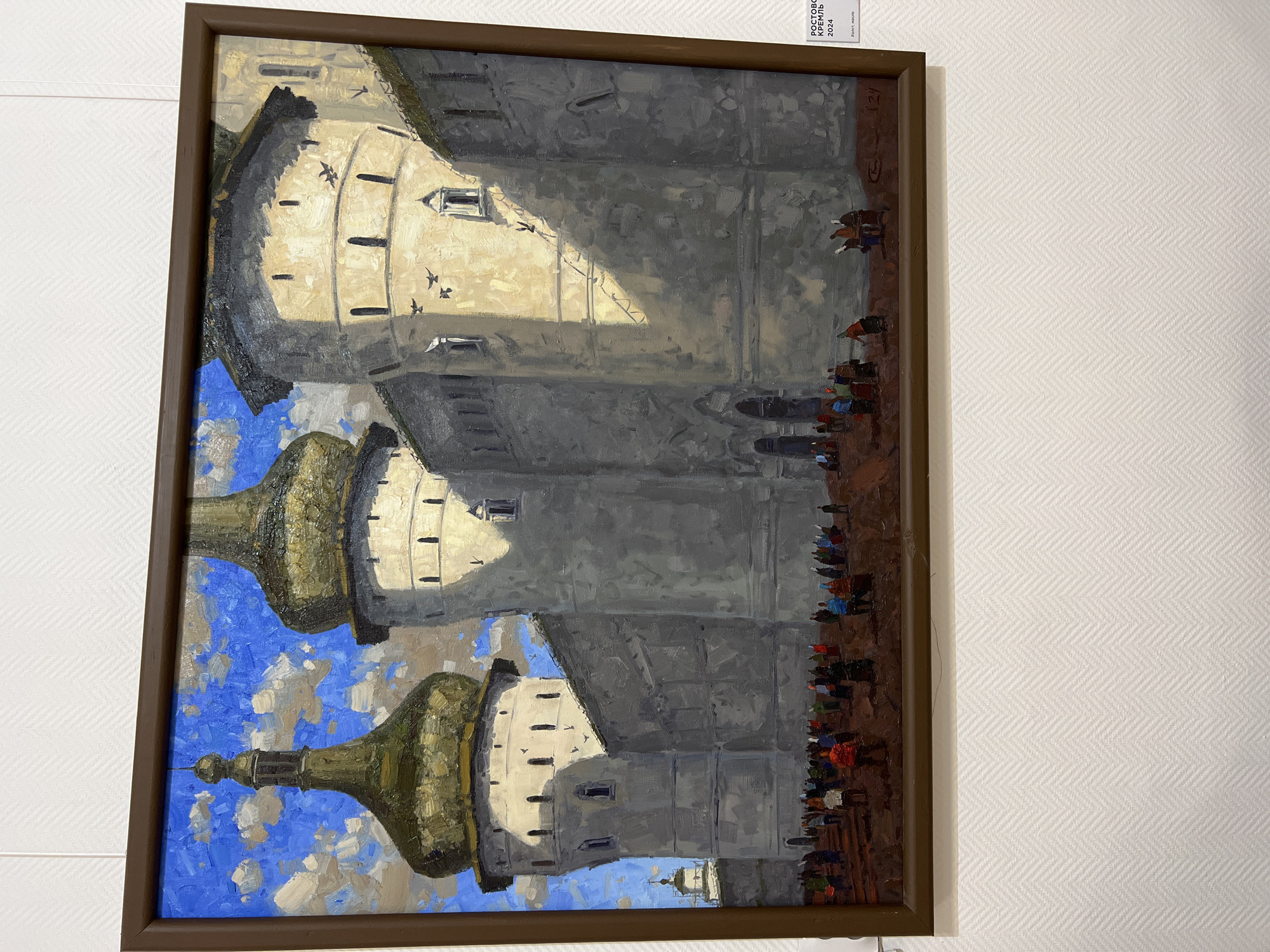 This is a painting of the Rostov Kremlin, a historic fortress in Rostov, Russia, depicting a sunny day with a bright blue sky filled with white clouds. The fortress walls are massive and grey, with several imposing towers topped with golden domes. One of the towers has a smaller, spired dome. Small birds are visible flying in the sky and near the towers. The foreground shows a crowd of people in colorful attire gathered on what appears to be a reddish-brown ground, possibly a courtyard or plaza. They are engaged in various activities, suggesting a public gathering or market. The overall scene is rendered in a painterly style with visible brushstrokes. A small white label is affixed to the right side of the painting's frame, indicating "РОСТОВ КРЕМЛЬ 2024" and below it, "ХУДОЖ. МАСЛО" (Artist. Oil). The painting is displayed on a wall with a white, textured pattern. There is no indication of the city of Novosibirsk in the image.
