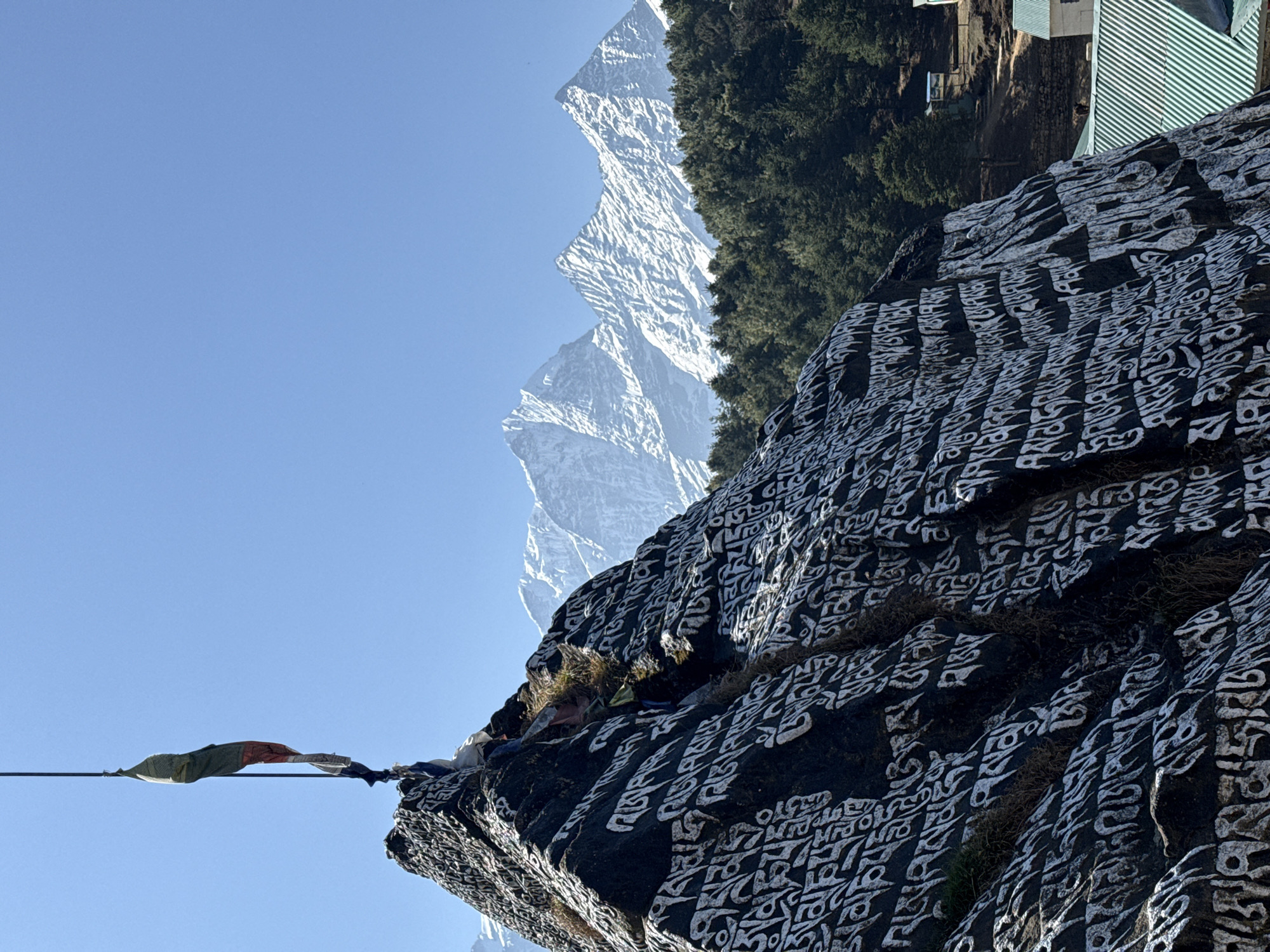This image captures a serene and majestic Himalayan landscape, set near Namche, Nepal. The foreground is dominated by a large, dark rock face, intricately covered with white inscriptions in what appears to be Tibetan script, likely mantras. These inscriptions cover the rock's uneven surface, creating a striking contrast.
Against a vibrant, clear blue sky, a single multi-colored prayer flag (green and red being prominent) is visible on the left, suspended horizontally on a wire.
In the mid-ground, a series of towering snow-capped mountains stretch across the horizon, their jagged peaks and ridges reflecting the bright sunlight. Below these impressive peaks, a dense forest of dark green evergreen trees covers the lower slopes. Towards the right, partially obscured by the rock and trees, are glimpses of a few small buildings with light-colored, possibly corrugated metal, roofs, indicating human settlement in this high-altitude region.
The time of day appears to be bright daylight, with strong sunlight illuminating the scene, casting clear shadows and highlighting the snow. The weather is clear and sunny with excellent visibility. The combination of the inscribed rock (Mani stone/rock), prayer flags, and snow-capped mountains firmly places the scene within the cultural and geographical context of the Himalayas, specifically consistent with the area around Namche. The image evokes a sense of peace and spiritual reverence amidst the grandeur of nature.
