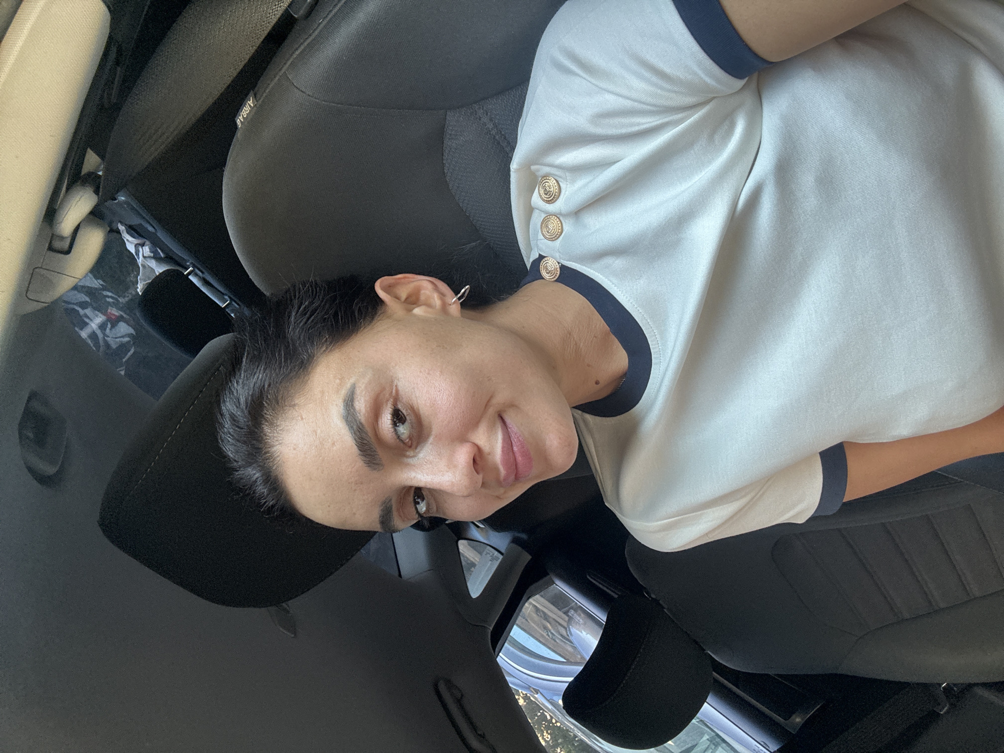 A woman with dark hair and eyebrows smiles as she sits in the passenger seat of a car. She is wearing a white t-shirt with a navy blue collar and trim, and a navy blue stripe on the sleeve. Two decorative gold buttons are visible on her left shoulder. A silver hoop earring is in her left ear. The interior of the car, including black seats and a seatbelt, is visible. The background shows a glimpse of the exterior through the car window, including what appears to be other vehicles and foliage. The lighting suggests it is daytime. The image is a selfie taken by the woman. The location is not specified in the image, but the prompt asks to assume it is Tbilisi, Georgia.