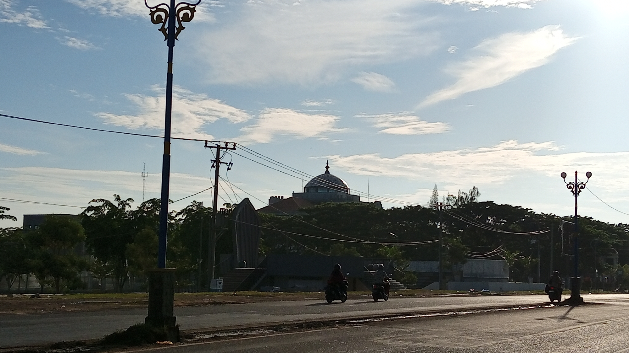 A daytime, eye-level, medium shot captures a street scene in Bireuen, Indonesia, under a bright, partly cloudy sky. A prominent, ornate street lamp with two lights stands on the left side of the frame, casting a shadow onto the asphalt road. In the middle ground, a domed building with a spire is visible above a dense line of trees, partially obscured by power lines and utility poles. Two individuals on motorcycles are depicted traveling on the road, one in the left lane and the other further back in the right lane. The road surface appears wet in places, reflecting the sky and surrounding objects. To the left of the street lamp, a dark, multi-story building can be seen. The overall environment suggests a developed urban or suburban area with a mix of infrastructure and greenery.