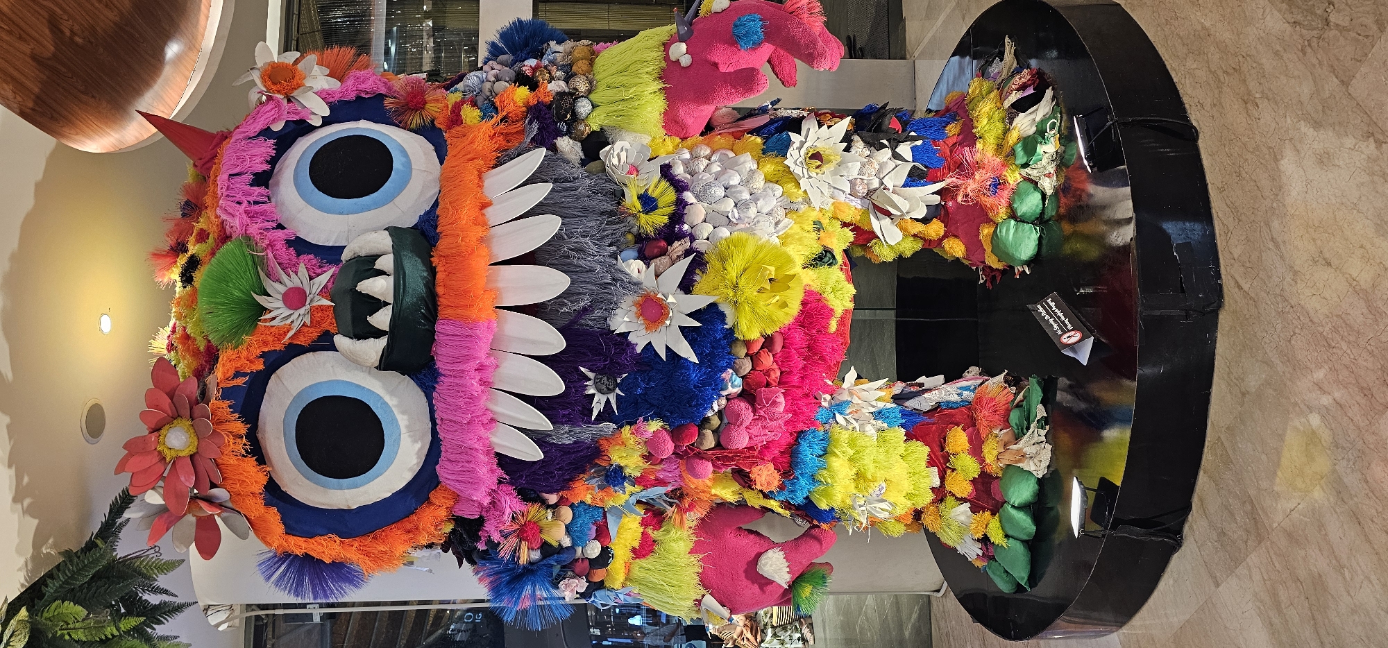 A large, colorful, vertically-oriented artistic display or sculpture stands indoors. It features oversized eyes with blue irises, a red horn, and a mouth with white teeth. The sculpture is covered in a variety of colorful materials, including yarn, fabric, and flowers, creating a textured and vibrant surface. It is positioned on a black circular base. The background includes interior architectural details such as walls, floors, and a section of the ceiling with a curved wooden panel. The sculpture is located in the city of Denpasar, Indonesia.