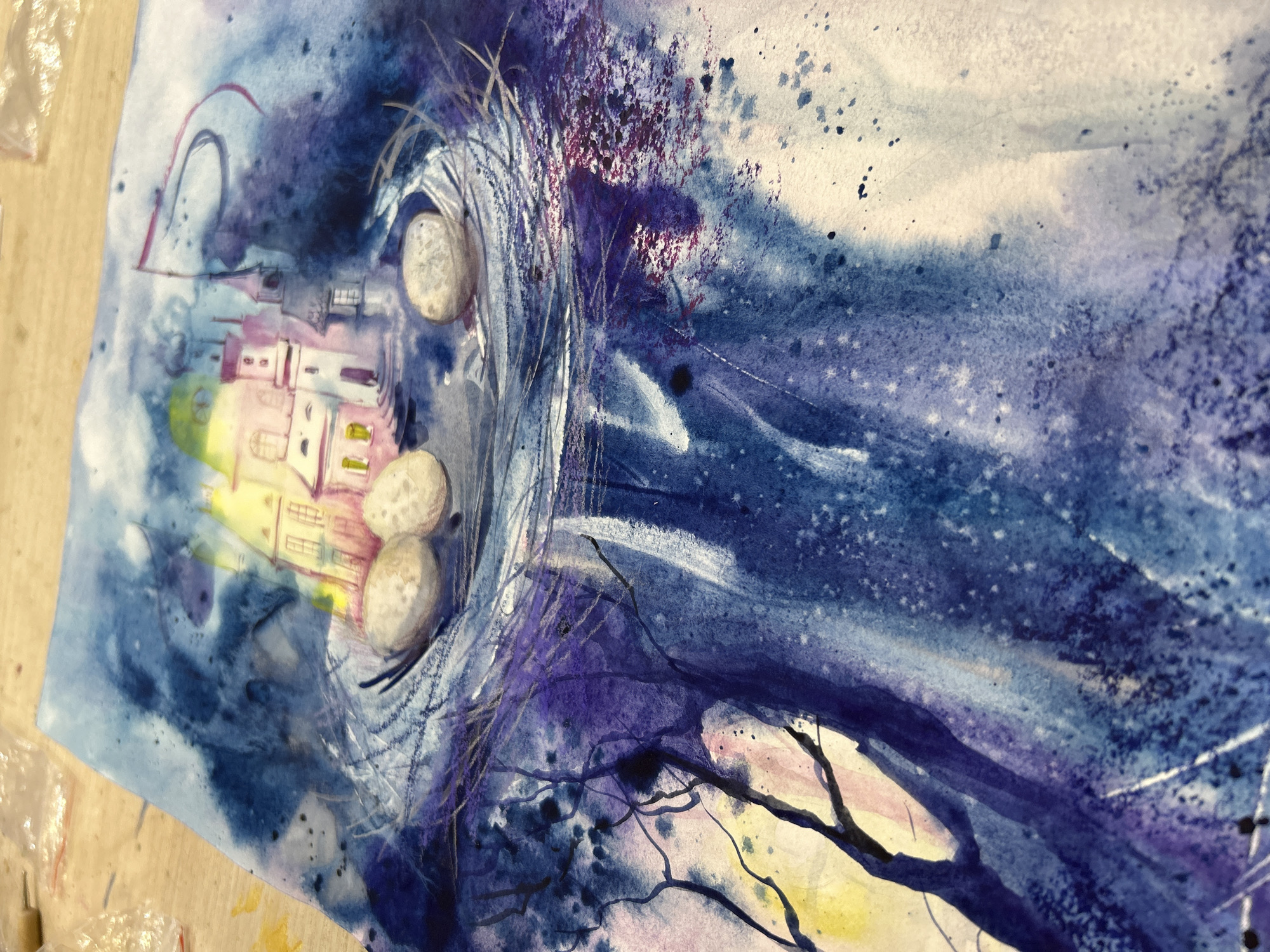 This is a photograph of a vibrant watercolor painting, dominated by a rich palette of deep blues and purples, accented with yellow, pink, and white. The artwork depicts a fantastical or dreamlike scene, likely set at night or twilight, characterized by dynamic washes and expressive splatters of paint.
In the upper-left area of the painting, a cluster of pastel-colored buildings, resembling a small town or castle, appears to glow with yellow and pink light from within its windows. Adjacent to these structures, three prominent oval-shaped objects, possibly eggs or smooth stones, are nestled within a swirling, protective element composed of intricate lines and lighter strokes, suggesting a nest or a celestial cradle.
The background is filled with expansive washes of indigo and violet, blending into lighter blues and whites, creating an ethereal atmosphere. Scattered dark specks across the canvas resemble stars or a spray of paint, adding texture and depth. Streaks of white and lighter blue suggest dynamic movement, perhaps of wind, clouds, or cosmic elements. From the lower-right portion, dark, intricate branches or roots extend into the turbulent background, providing a stark contrast and grounding the fantastical scene.
The overall impression is one of mystery, wonder, and a sense of coziness or protection around the glowing town. The painting is shown resting on a light wooden surface, with a small amount of clear plastic packaging visible on the left side of the frame, outside the artwork itself. There is no visible text in the image.
