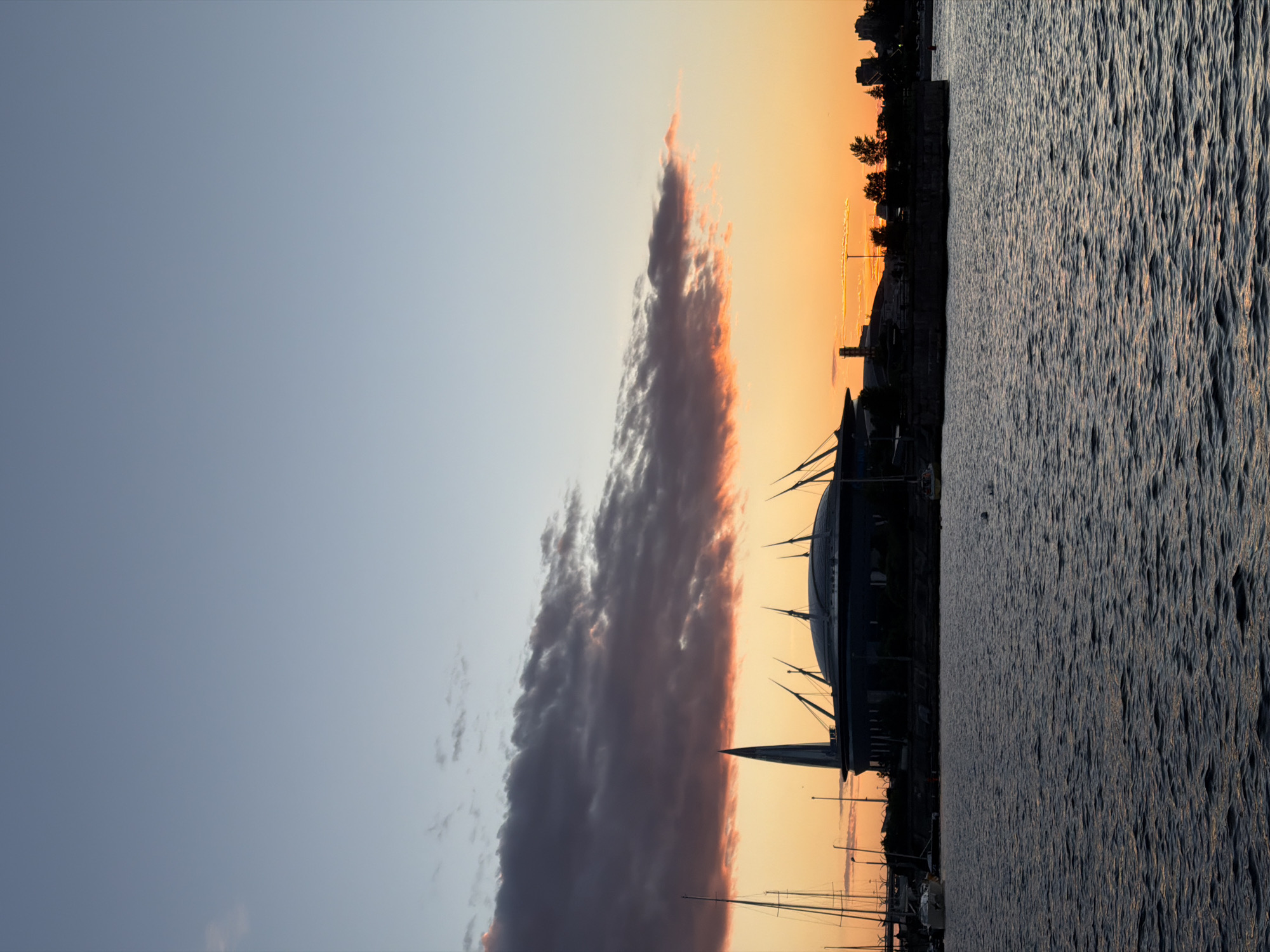 This is a vertical, eye-level, medium shot of the Gazprom Arena stadium in Saint Petersburg, Russia, during a sunset. The stadium is silhouetted against a sky that transitions from a clear pale blue at the top to hues of orange and pink near the horizon. Dark, heavy clouds with tinges of pink and orange are positioned above the stadium, obscuring the sun. The stadium itself is a dark, indistinct shape with its characteristic pointed spire and canopy structure visible. In the foreground, a dark body of water, likely the Neva River or a surrounding canal, reflects the dim light of the sunset. Several sailboats with tall masts are moored to the left of the stadium, their silhouettes also dark against the sky. Trees and other indistinct structures are silhouetted along the embankment behind the stadium. The overall scene is serene and captures the architectural landmark during twilight.