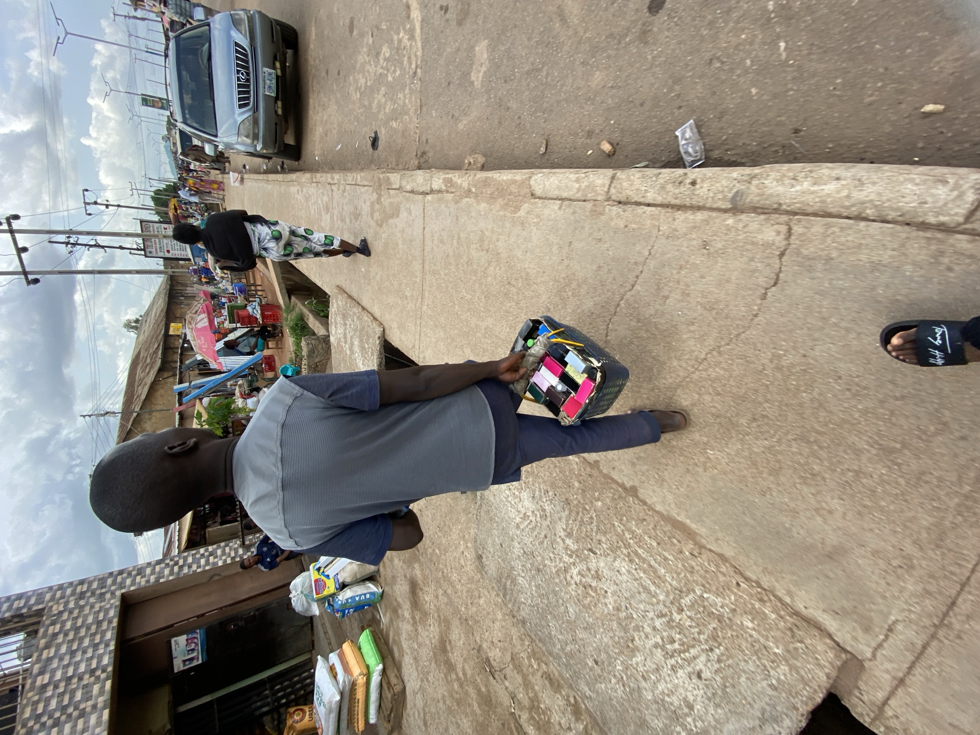 A medium shot captures an individual from behind, walking on a sidewalk alongside a bustling street in Bukuru, Nigeria. The person is wearing a blue shirt with gray sleeves, dark pants, and is carrying a box filled with various small items.
To the left, a building and bags of fertilizer or other product can be seen along with a vendor’s storefront. The sidewalks are unkempt and show their age through cracks and weathering. To the right, part of a foot wearing a black sandal can be seen.
In the background, there's a clear view of the street. A silver van is parked on the side, and a person clad in a black top and patterned trousers walks away further down the street. Market stalls, people, and other vehicles suggest a busy trading area.
The sky is partly cloudy, with power lines overhead, typical of an urban environment. Overall, the image portrays a common street scene in Bukuru, displaying the everyday life and commerce of the area.