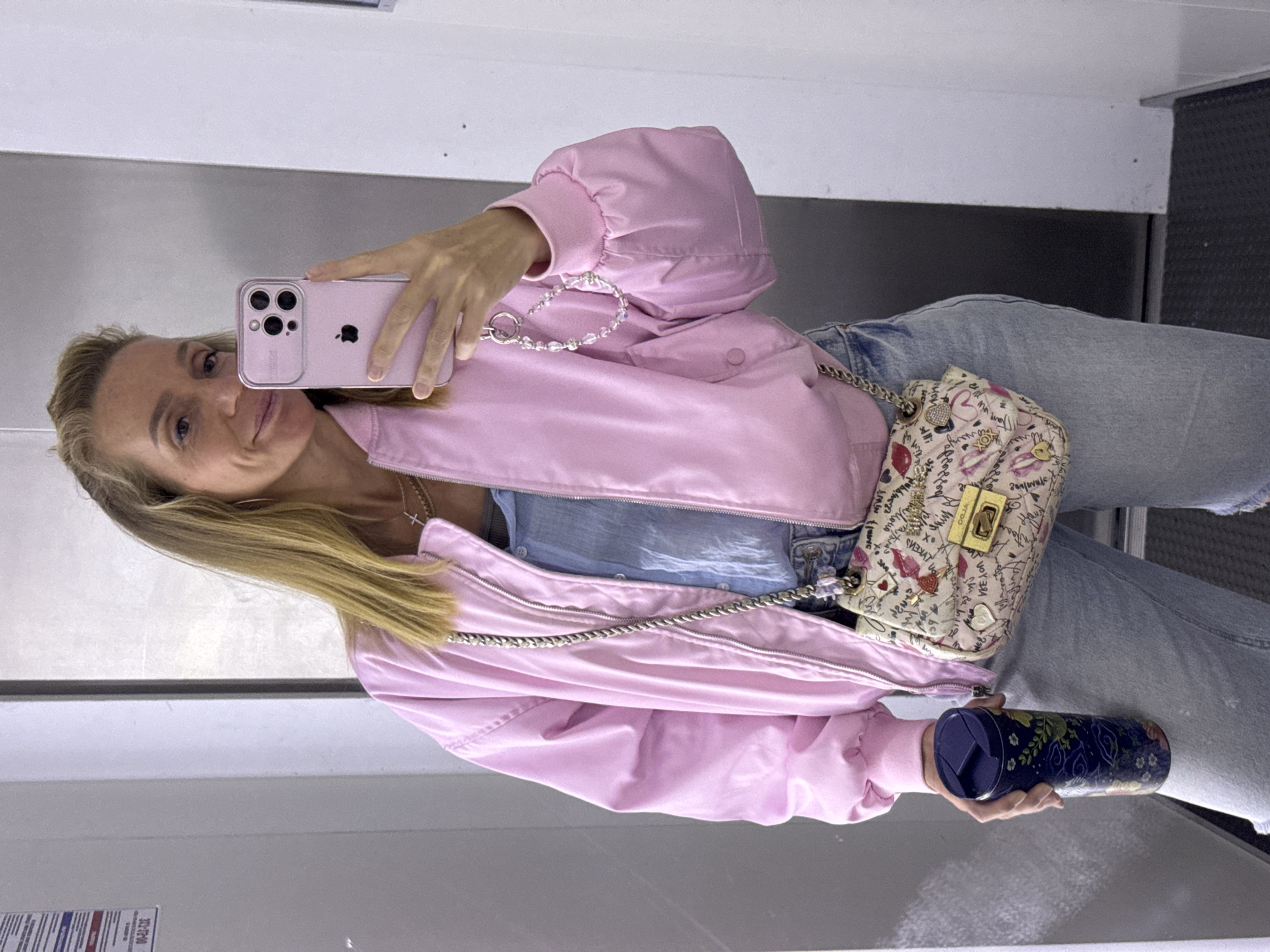An unidentified woman is standing in an elevator and taking a selfie. She is wearing a pink bomber jacket, a blue top, and light wash jeans. She has a small, patterned crossbody bag with a gold clasp and a silver chain strap. She is holding a dark blue travel mug in her left hand and her phone in her right hand. Her hair is blonde and pulled back. The elevator walls are light gray and white. There is a sign on the left wall of the elevator.