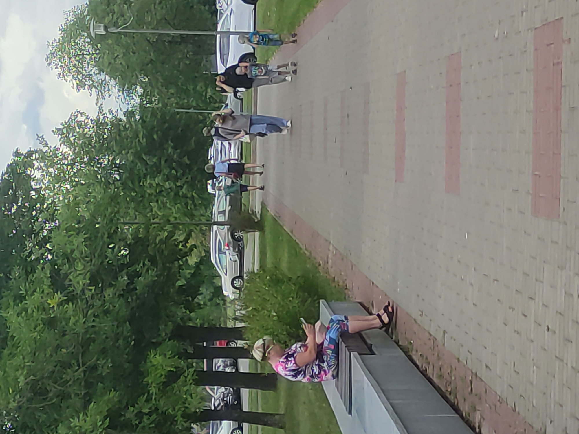 A woman with short blonde hair, wearing a brightly colored floral print t-shirt and patterned leggings, sits on a long concrete bench in a park, looking at her phone. She is wearing dark sandals. The bench is adjacent to a paved walkway with red brick inlays. In the background, several people are walking along the path, including a family with two young boys. Trees line the walkway and a parking lot with several cars is visible further in the background. The setting appears to be a public park in the city of Mahilyow, Belarus.