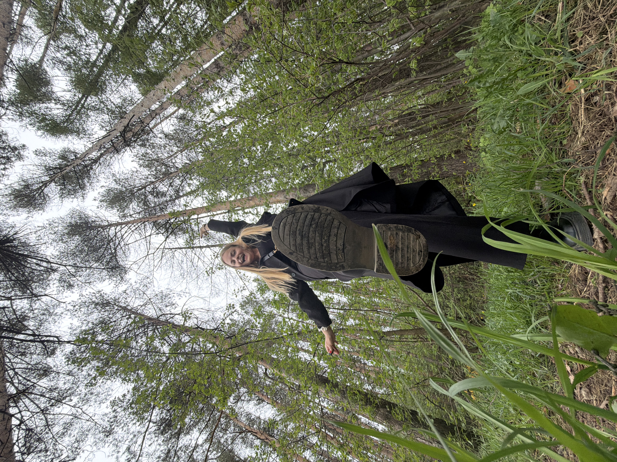 A woman with long blonde hair, wearing a black coat and shoes, is standing in a forest. The view is from below, looking up towards her. The woman is smiling and has one arm outstretched. The sole of one of her shoes is visible and prominent in the foreground. Tall trees surround her, and green foliage is visible. The specific location in Izhevsk, Russia, cannot be determined from this image.