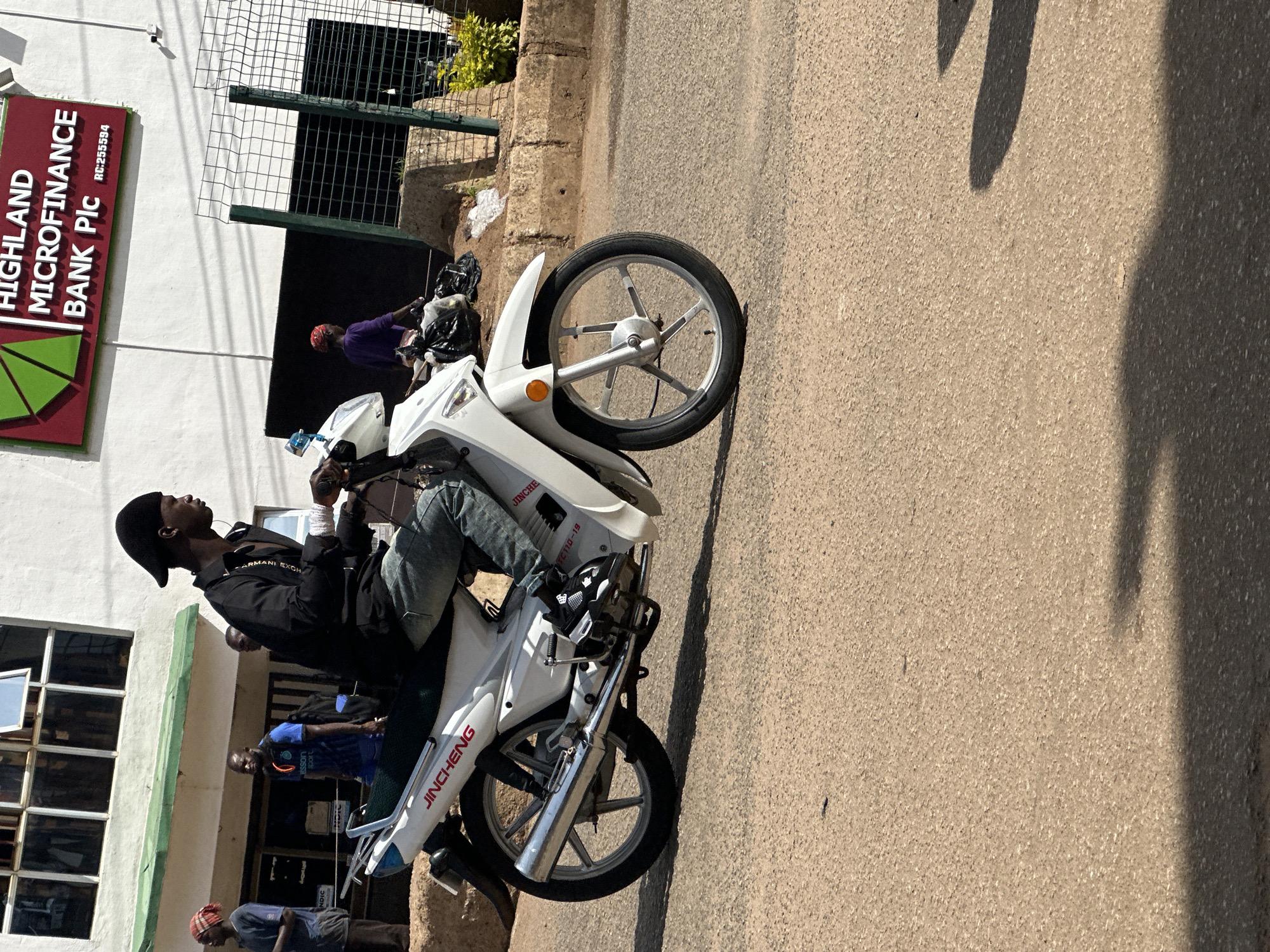 A man wearing a black jacket, blue jeans, and a black cap is sitting on a white motorcycle. He is looking to the side with a neutral expression. The motorcycle has the brand name "JINCHENG" written on its side. In the background, there is a building with a sign that reads "HIGHLAND MICROFINANCE BANK Plc RC:255594". Other people are visible in the background, some are standing, and one is wearing a purple shirt. The scene appears to be on a street or in front of a building. The lighting suggests it is daytime, and the weather seems to be sunny, casting shadows on the ground. The overall setting is an urban environment in Bukuru, Nigeria. The image is oriented vertically.