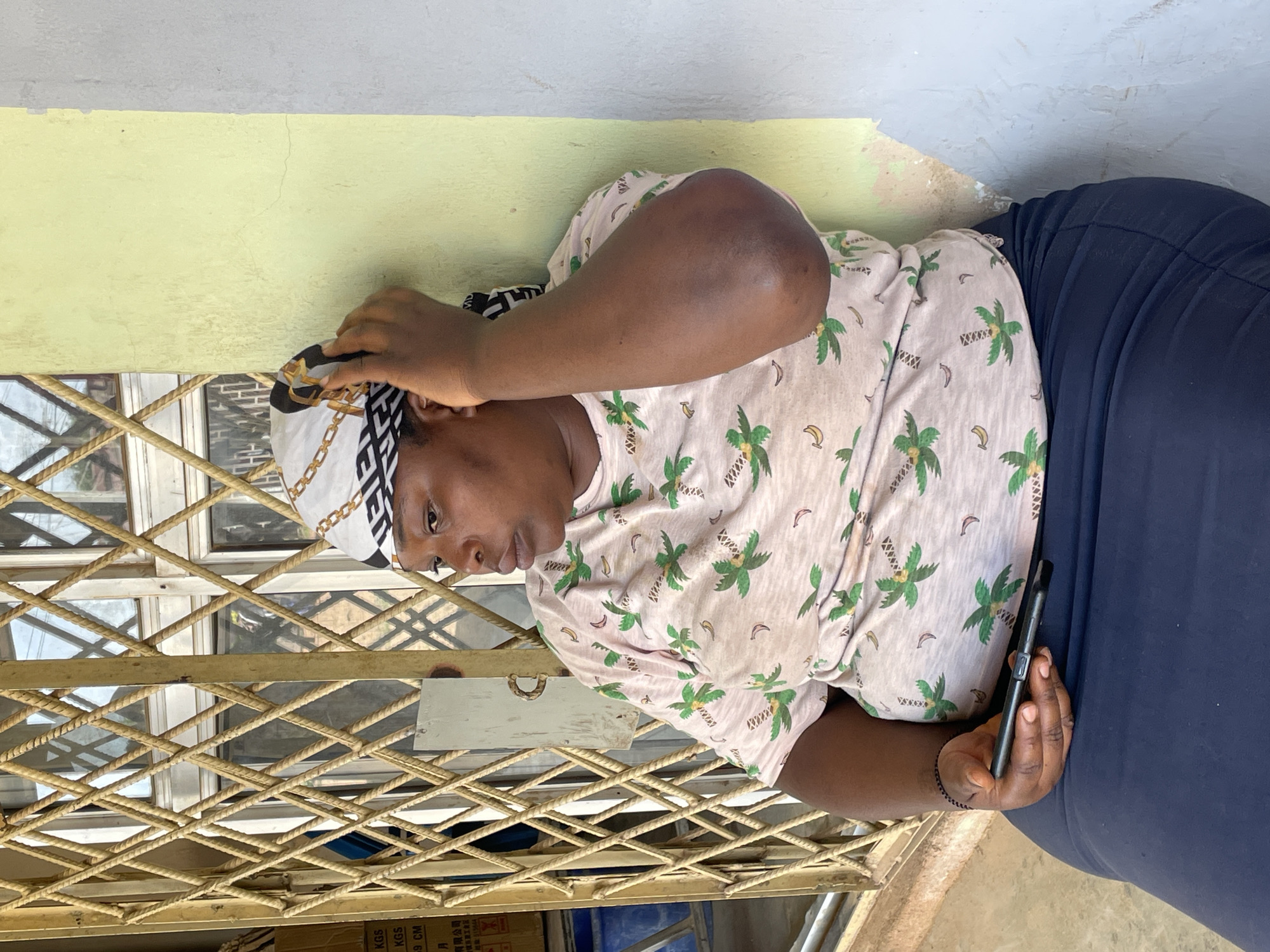 A medium shot captures a woman sitting outdoors, leaning against a pale green wall. She is wearing a light pink t-shirt adorned with a pattern of green palm trees and yellow bananas, and a dark blue skirt. A white headscarf with a black and gold geometric pattern covers her hair. Her right hand is raised to her head, adjusting the scarf, while her left hand rests on her lap, holding a black smartphone. She is looking away from the camera. Behind her, a diamond-patterned metal gate is visible against a window. The environment suggests an urban setting in Barakin Akawo, Nigeria, with the presence of a building exterior and a street-facing gate.