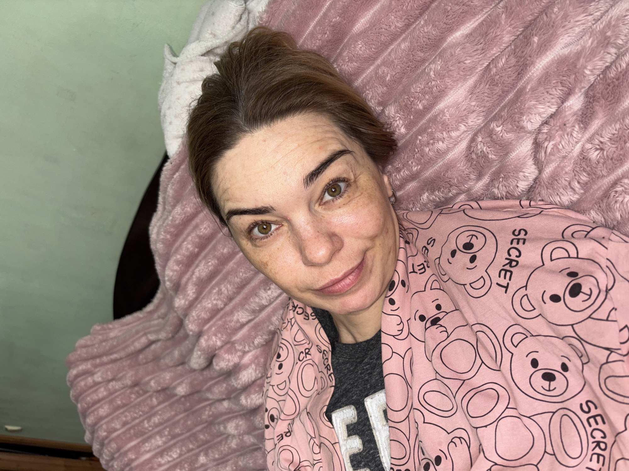 An individual is lying in bed, covered by a pink blanket with a teddy bear pattern. The person is looking directly at the camera with their head tilted. They are wearing a gray t-shirt under the blanket. The background shows a wall painted in a light green color, and a portion of a wooden headboard. The lighting appears to be artificial, suggesting an indoor setting. The scene is consistent with a bedroom in a residential dwelling in Murino, Russia, given the context.