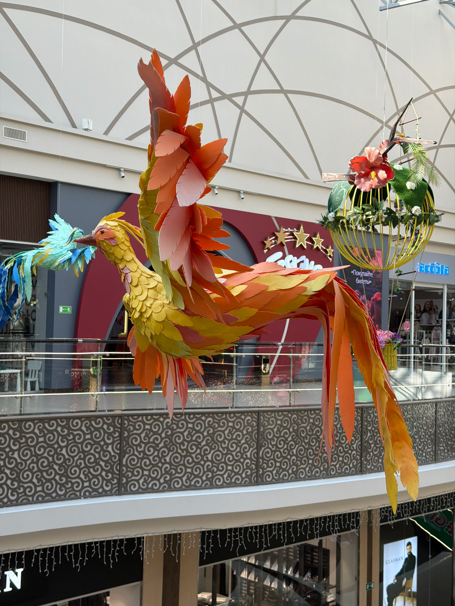 This is a full-shot, eye-level, medium-distance, indoor photograph of a large, colorful, artificial bird sculpture suspended from the ceiling of a shopping mall. The bird, which appears to be a phoenix, is made of paper or fabric and is predominantly yellow, orange, and red. It is depicted in mid-flight, with its wings spread and its head turned to the left, where another smaller, blue bird sculpture is visible. The background shows the interior of a multi-story shopping mall, with balconies, railings, storefronts, and people in the distance. The mall has a modern design with a white, curved ceiling. The sculptures are likely part of a seasonal or promotional display. This scene is located in the city of Pavlodar, Kazakhstan, within a shopping mall.