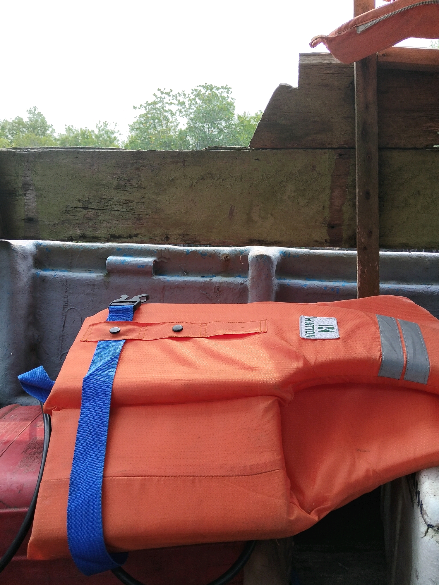 An orange life vest with blue straps is secured within a boat's interior. The life vest, with a white tag bearing the brand "HATTORI" and a green logo, is folded. The interior of the boat features worn, weathered wooden planks and a grey, textured surface with blue paint. Outside the boat, blurred green trees and a bright white sky are visible. The scene is set in Kembung Luar, Indonesia.