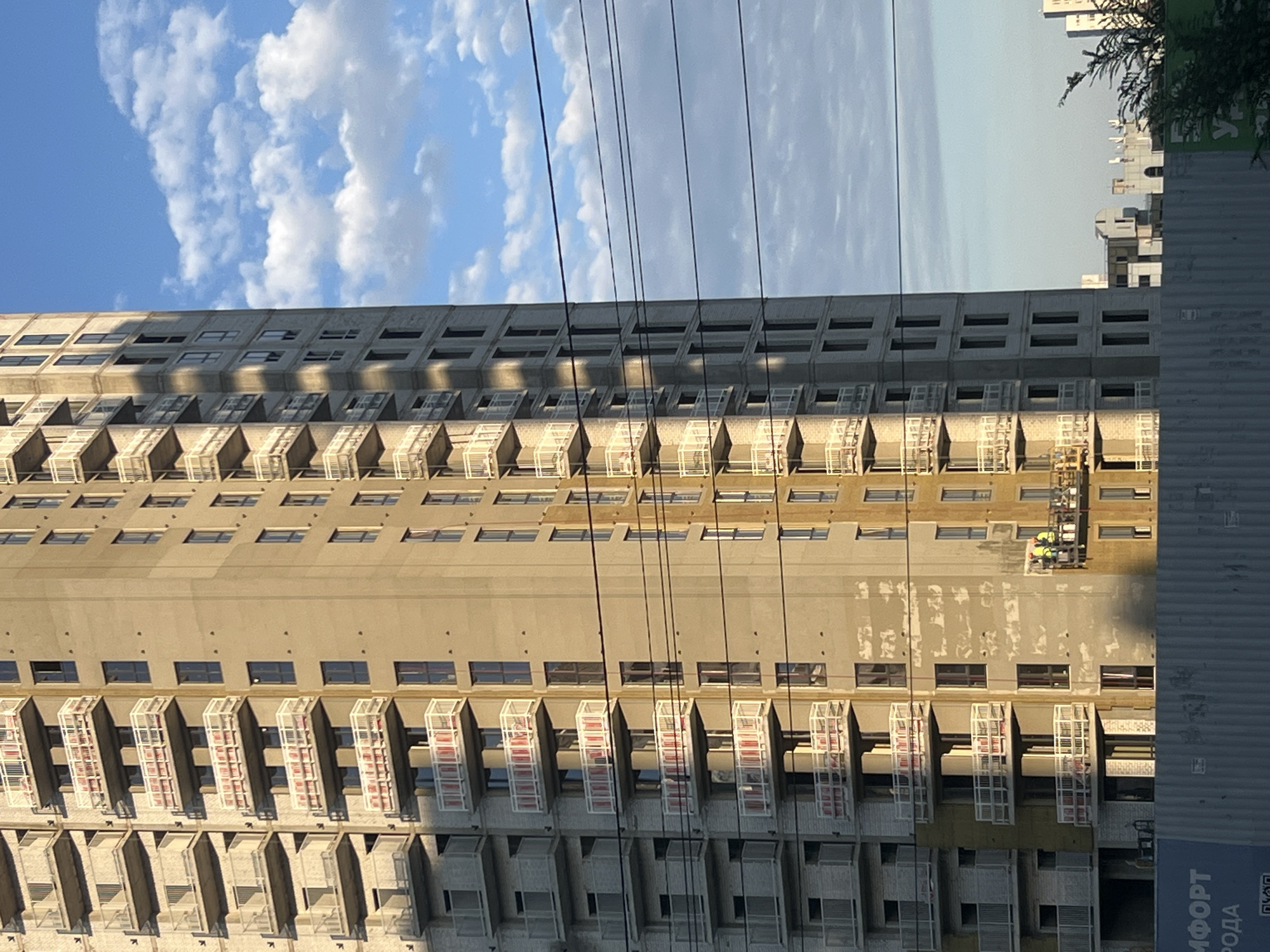 The image shows a high-rise residential building under construction in Volgograd, Russia. The building is tall and narrow, with many balconies. The facade is made of concrete and has a warm, sandy color. The sky is blue with white clouds. There are power lines in the foreground. Two construction workers are visible on a suspended platform on the side of the building, appearing to be engaged in work. In the lower left corner, a blue fence with some text in Russian is visible. In the distance, other buildings are partially visible. The lighting suggests it is either early morning or late afternoon, with the sun casting shadows on the building.