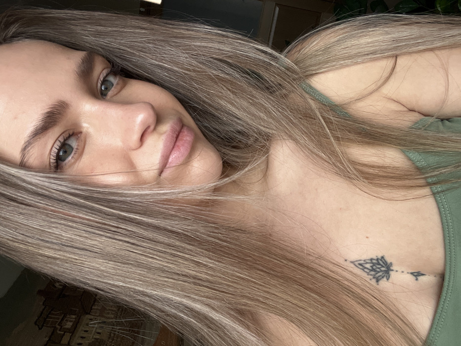 A close-up portrait shows a young adult female with long, straight, light blonde hair and blue-grey eyes, looking directly at the camera. She has a fair complexion and a subtle, closed-mouth smile. She is wearing a sage green tank top or camisole. A black linear tattoo, depicting a stylized lotus flower design with a descending line and a small geometric shape, is centered on her sternum, visible above the garment's neckline. The background is out of focus, indicating an indoor setting in Saint Petersburg, Russia. To the upper left, a dark, blurry object is visible, consistent with a tapestry or print depicting European architecture. To the right, a lighter, indistinct area with suggestions of interior furnishings and possibly foliage can be seen.