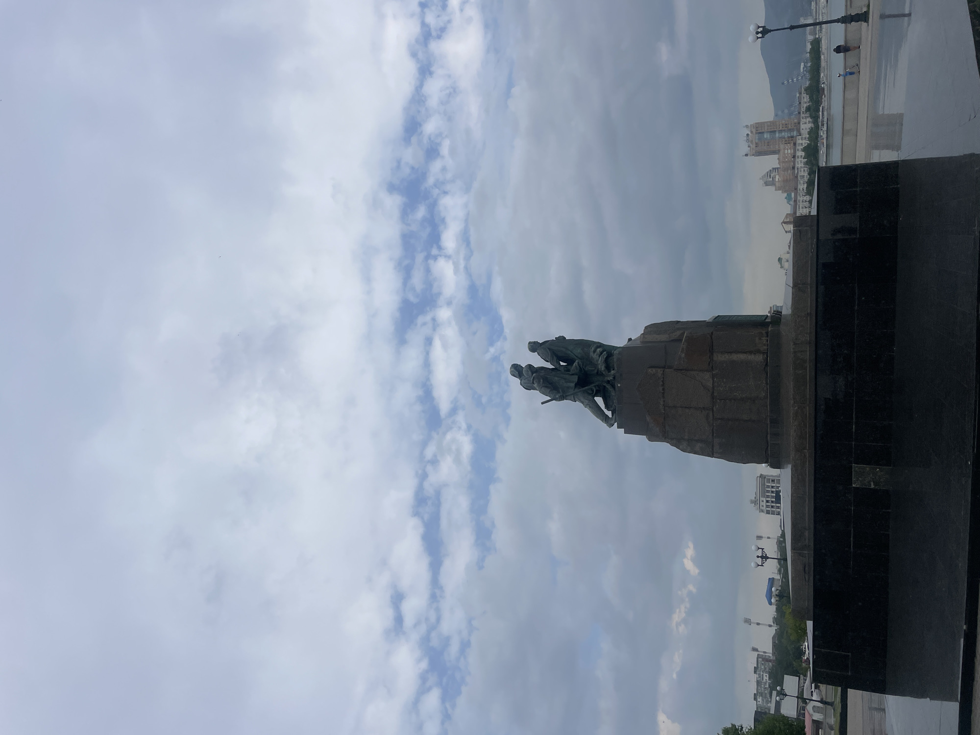 The image shows the Monument to the Heroes of the Civil War in Novorossiysk, Russia, a bronze sculpture depicting three figures in dynamic poses, standing on a granite pedestal. The monument is set against a backdrop of a cloudy sky. In the distance, a city skyline with buildings and a body of water can be seen. The foreground features a dark, reflective surface, possibly polished granite, which mirrors the sky and the monument. Several streetlights are visible, and a few people are present on a promenade by the water. The overall scene is overcast and somewhat somber.